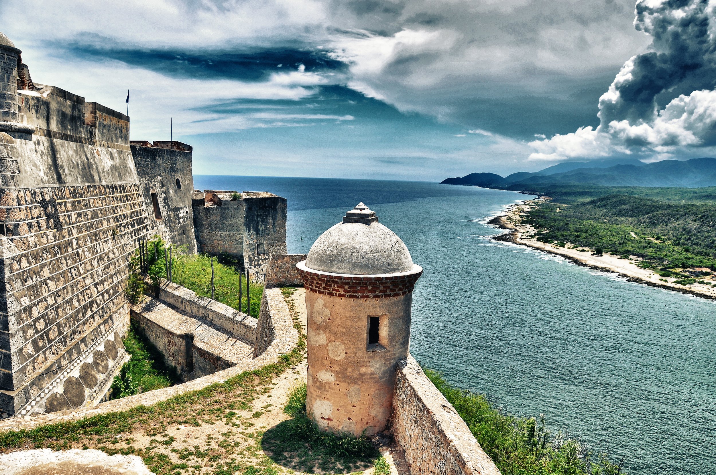 Fortress overlooking a coastal landscape with blue ocean and rugged shoreline, featuring clouds in the sky.
