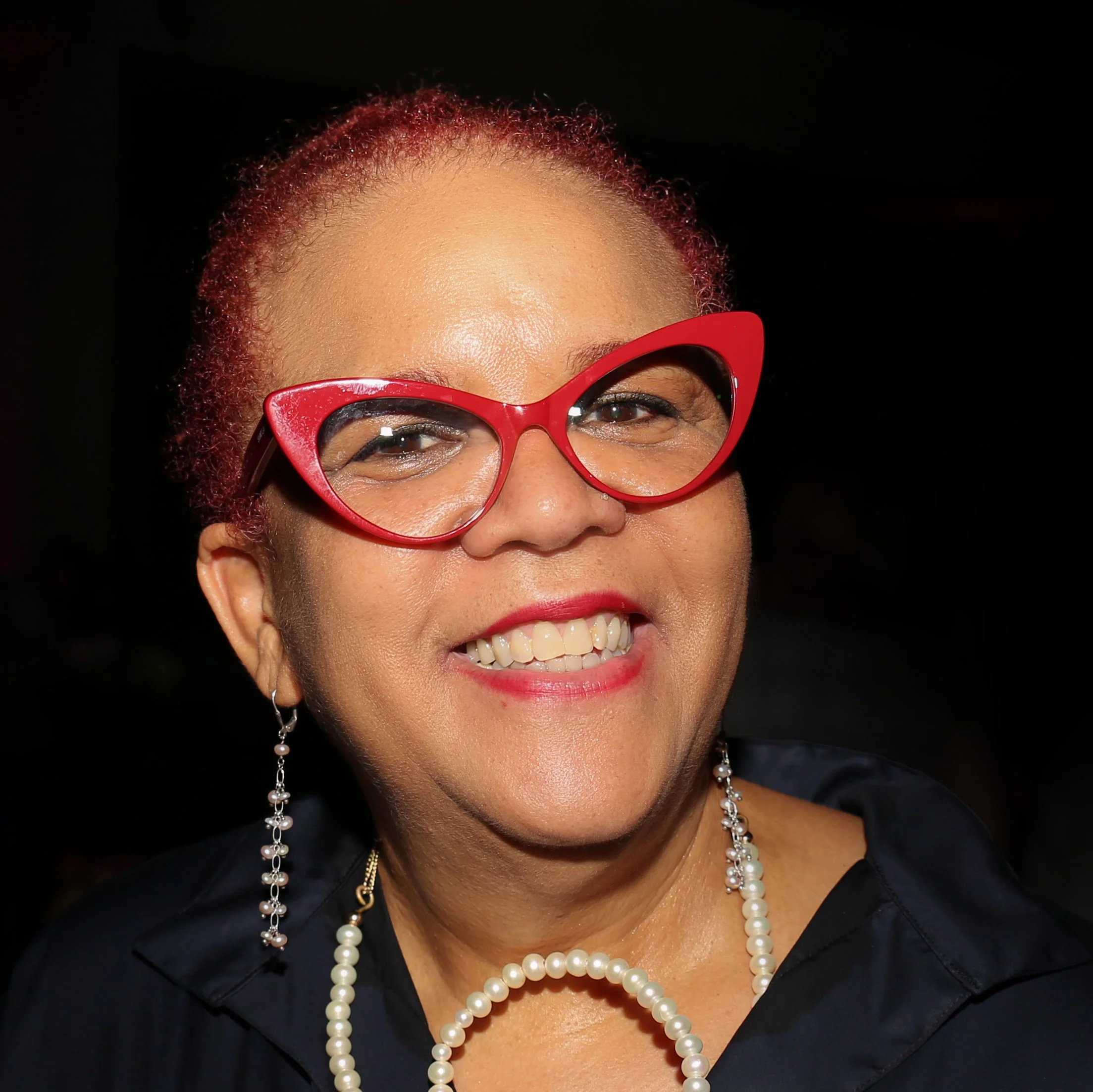 Person smiling with red glasses and red hair, wearing earrings and a pearl necklace.