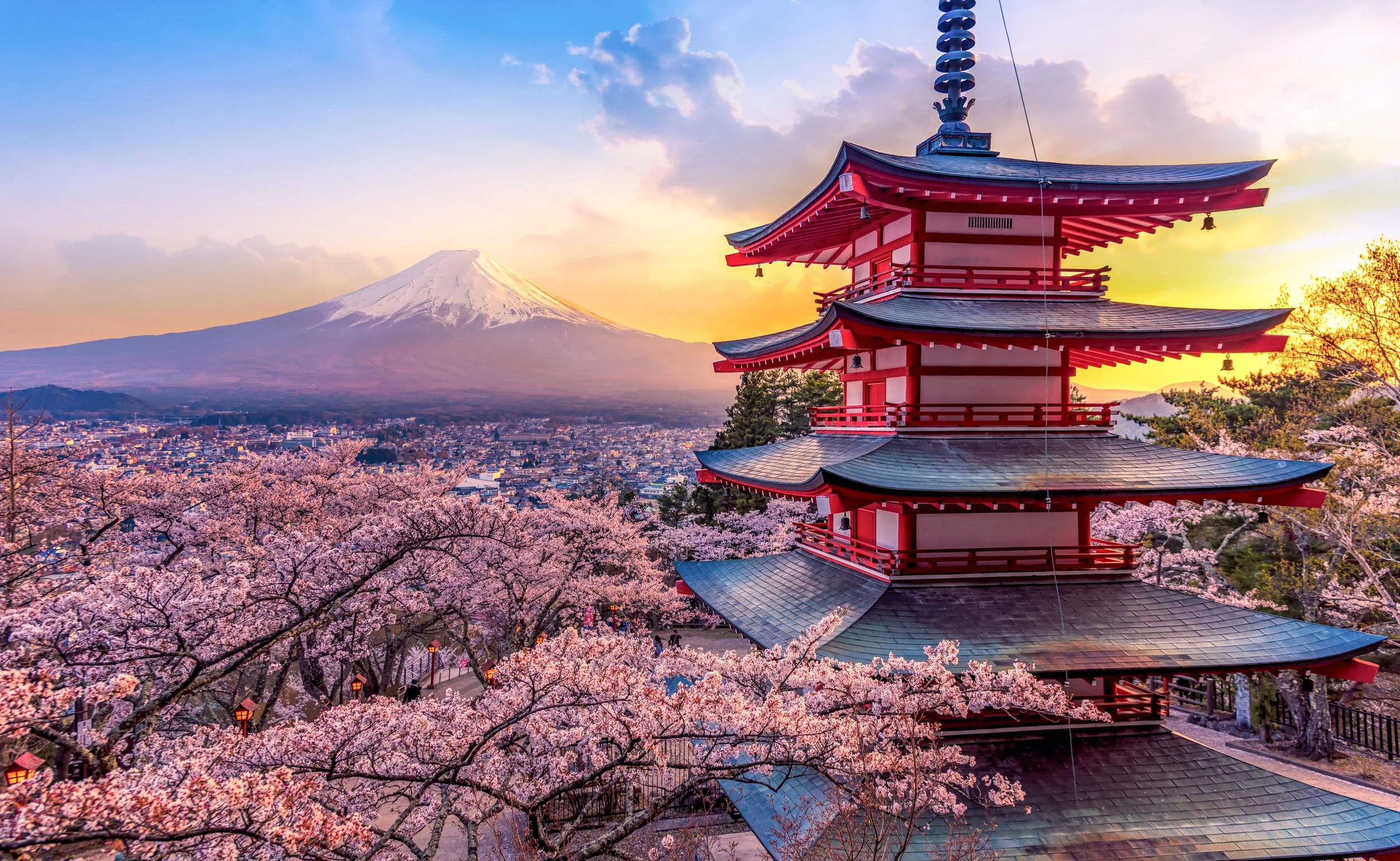 A traditional Japanese pagoda with multiple tiers in pink and black, surrounded by cherry blossoms in full bloom. In the background, Mount Fuji with snow-capped peak is visible during sunset or sunrise with a sky filled with cloudy and warm hues.