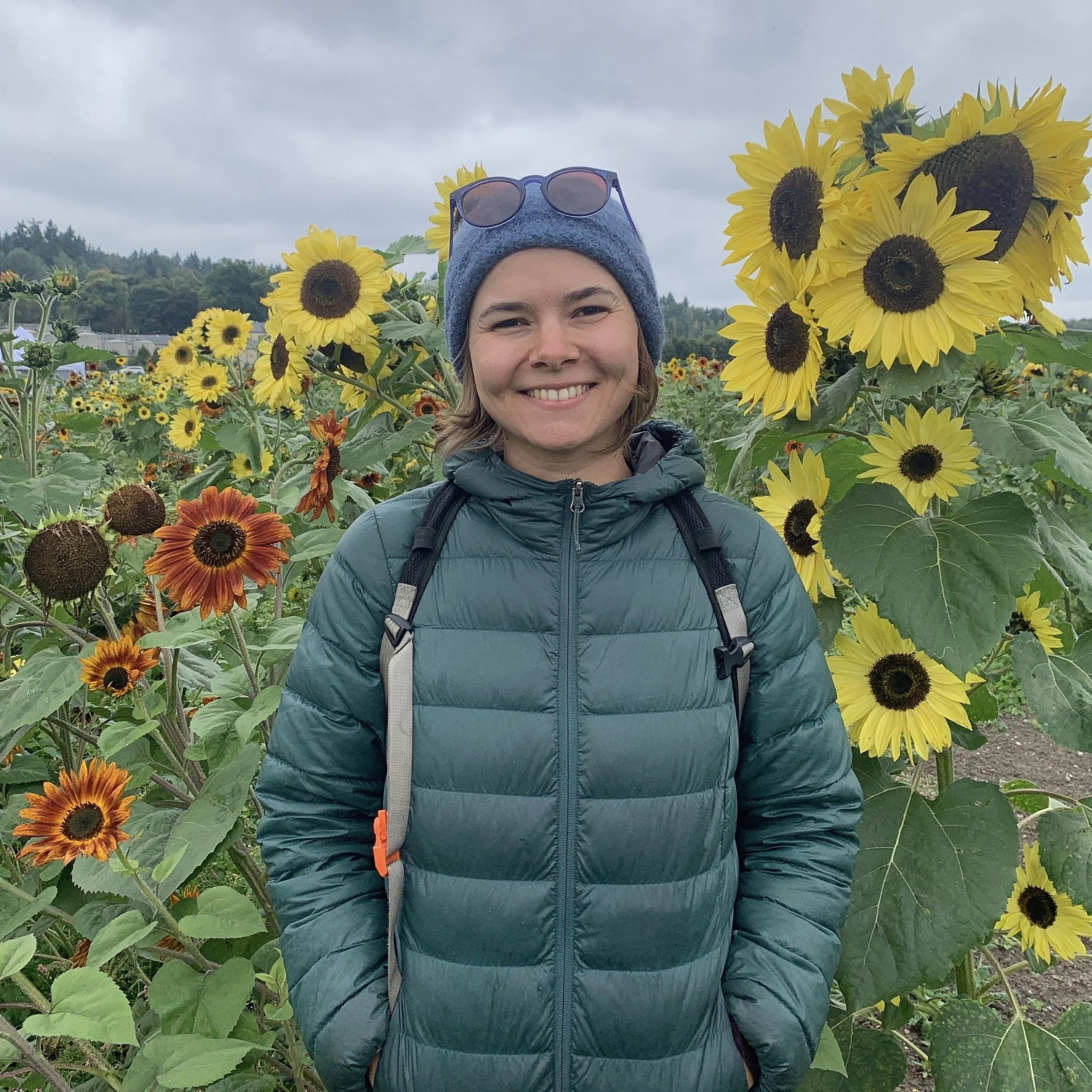 A woman smiling in a sunflower field, wearing a blue beanie, sunglasses, a teal puffer jacket, and carrying a backpack.