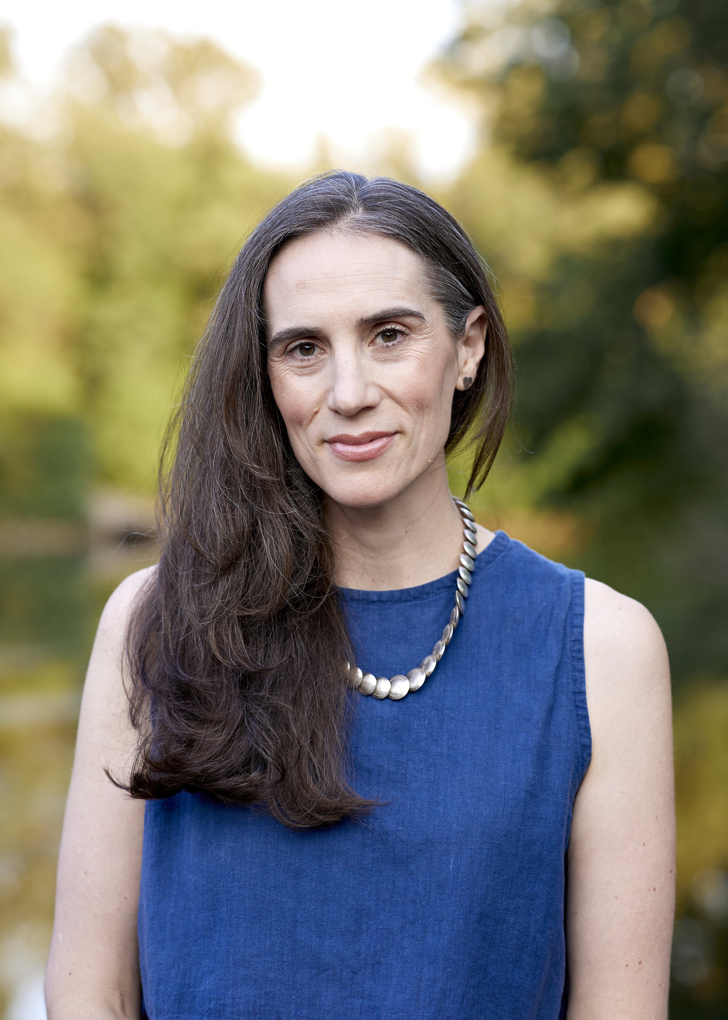 A woman with long brown hair wearing a blue sleeveless top and a pearl necklace, posing outdoors with a blurred greenery background.
