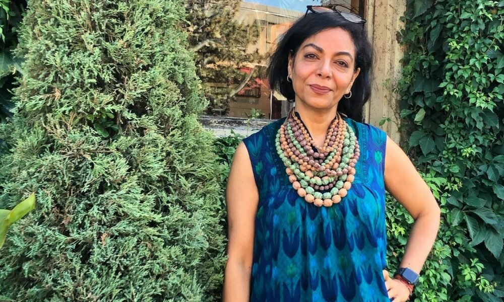 A woman standing next to a lush evergreen shrub, wearing a blue patterned sleeveless dress and multiple colorful beaded necklaces. She has dark hair and is smiling slightly, with one hand on her hip. The background includes greenery and part of a building.