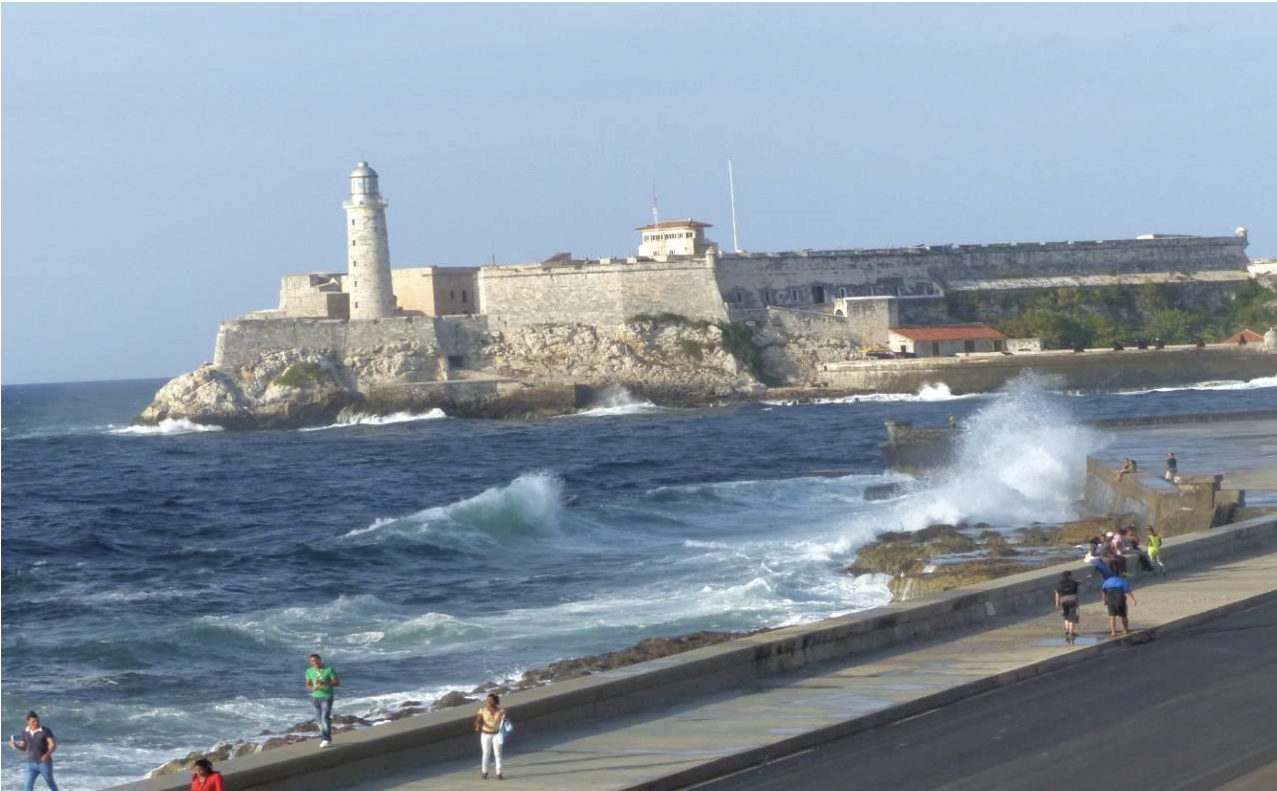 View of El Morro Fortress in Havana with ocean waves and people walking along the Malecón sea wall.
