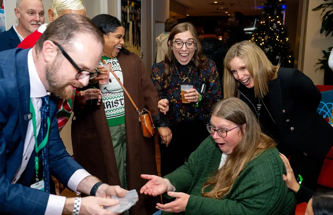 A group of people gathered around a woman with long hair in a green sweater, who appears surprised or excited, as a man in a blue plaid suit hands her a gift. The others are smiling and laughing in a festive setting with holiday decor.