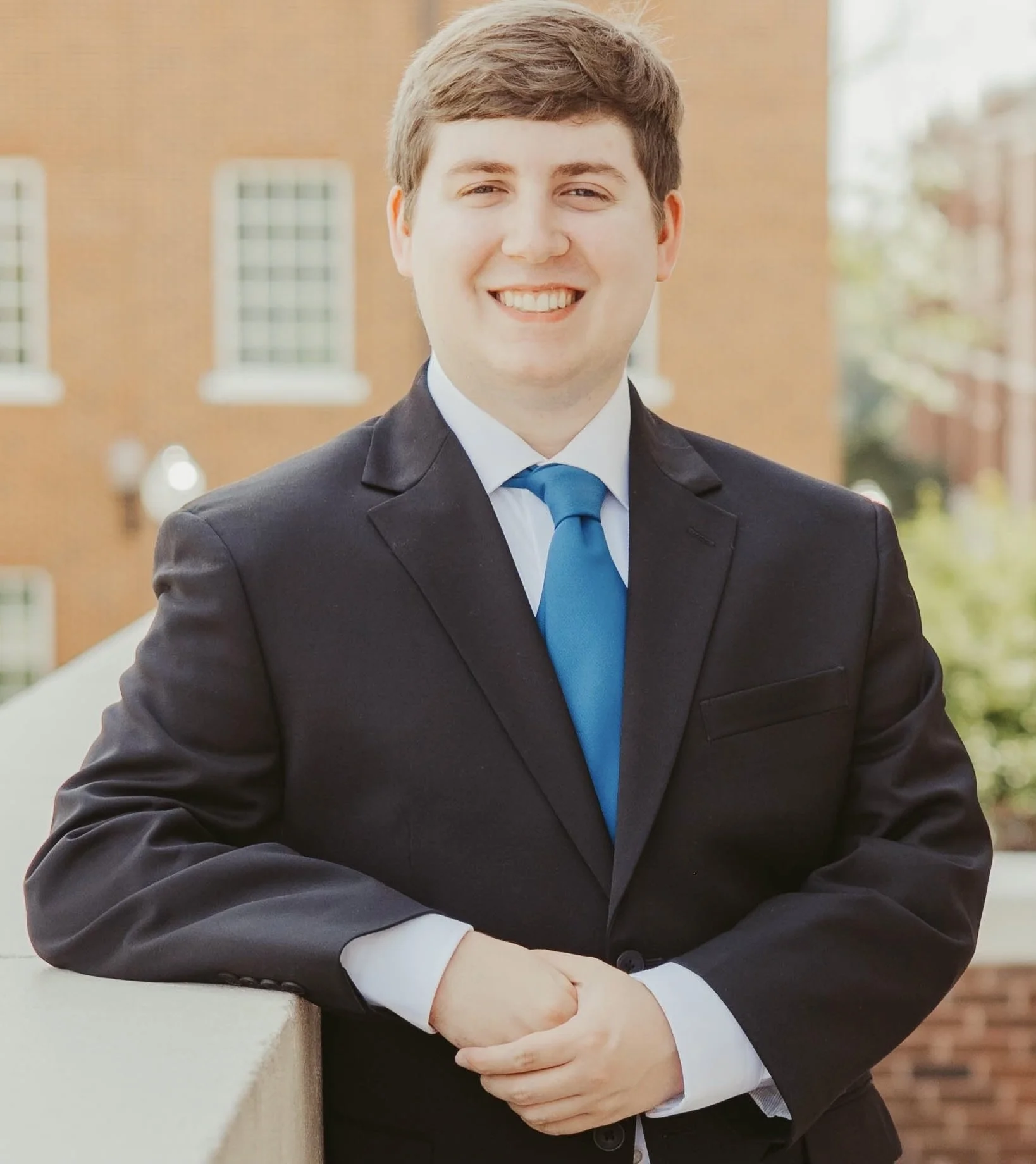 A young man in a black suit, white shirt, and blue tie, smiling outdoors with a brick building in the background.