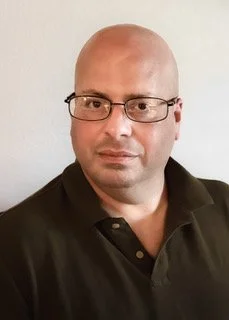 A smiling man wearing sunglasses and a blue checkered shirt.