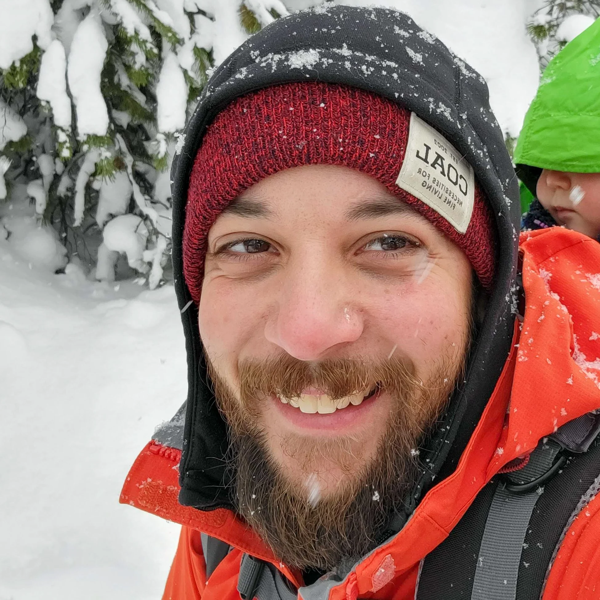 A smiling man with a beard dressed in winter gear, including a red knit cap, a black hood, and an orange jacket, is in a snowy outdoor setting with snow-covered trees in the background.