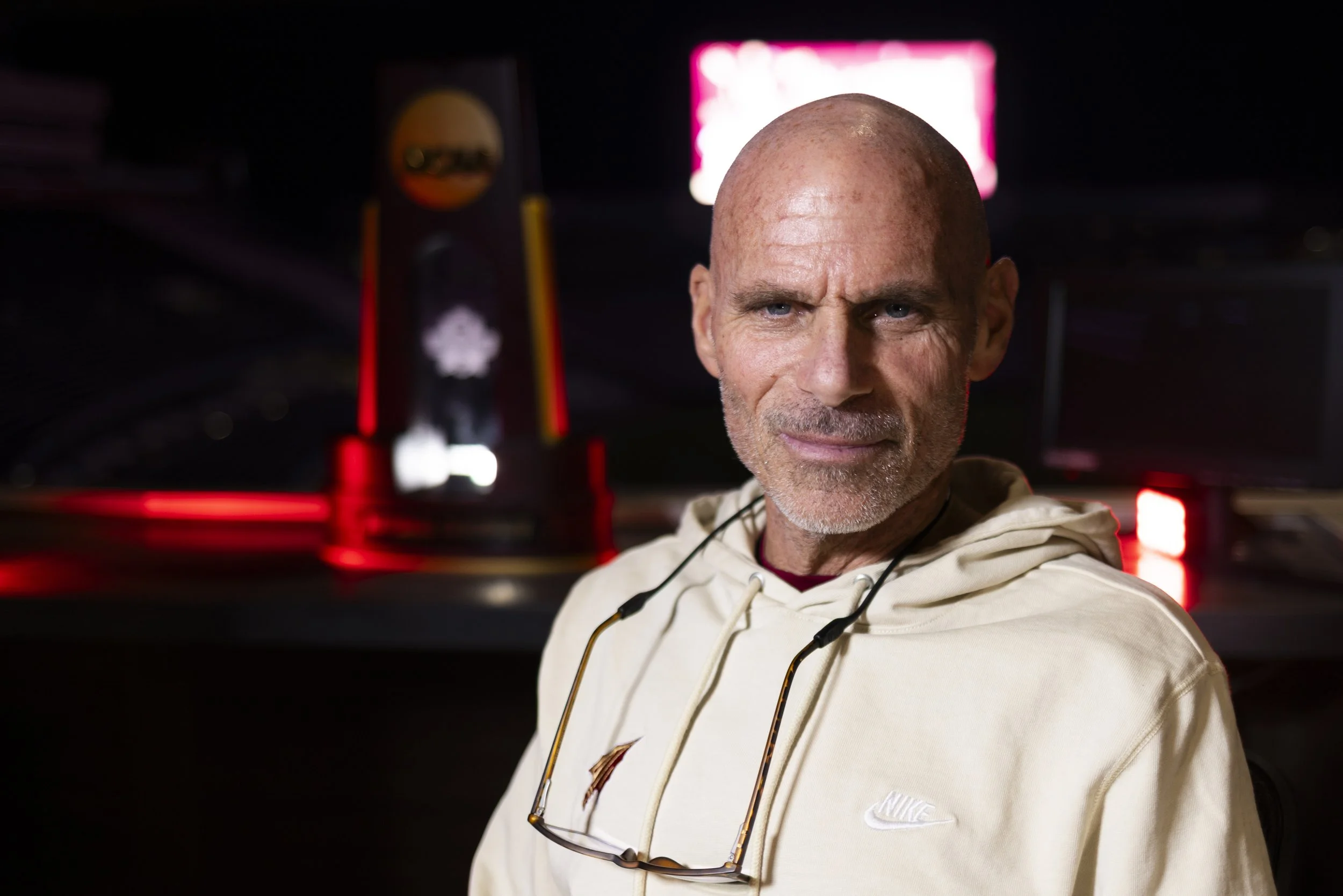 Close-up of a smiling man with a shaved head and light beard, wearing a Nike hoodie, with sports trophies and a digital sign in the background.