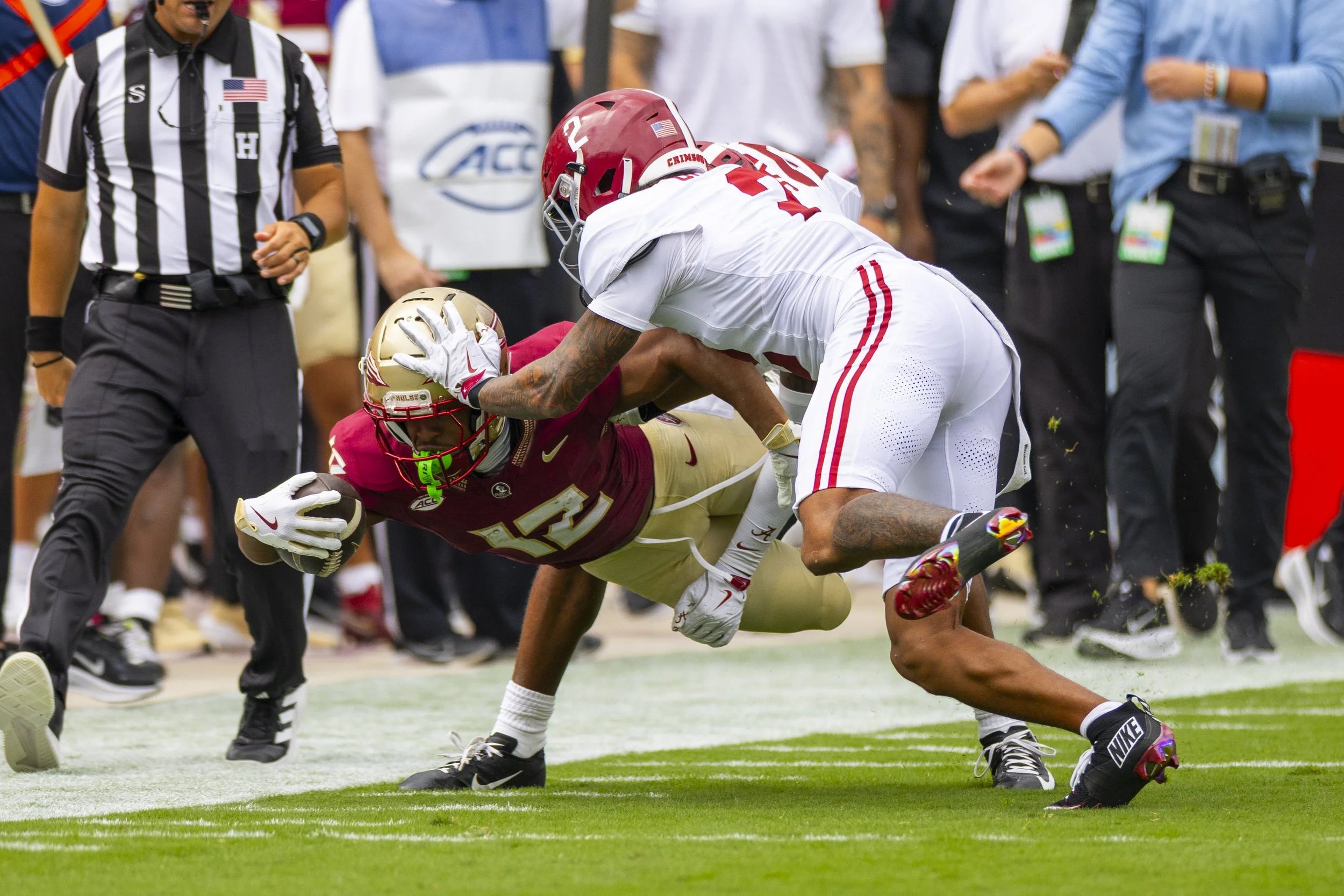 An intense American football game with a player from Florida State University being tackled by a player from the University of Alabama. The Florida State player is holding the football while on the ground, and the Alabama player is grabbing him aroun