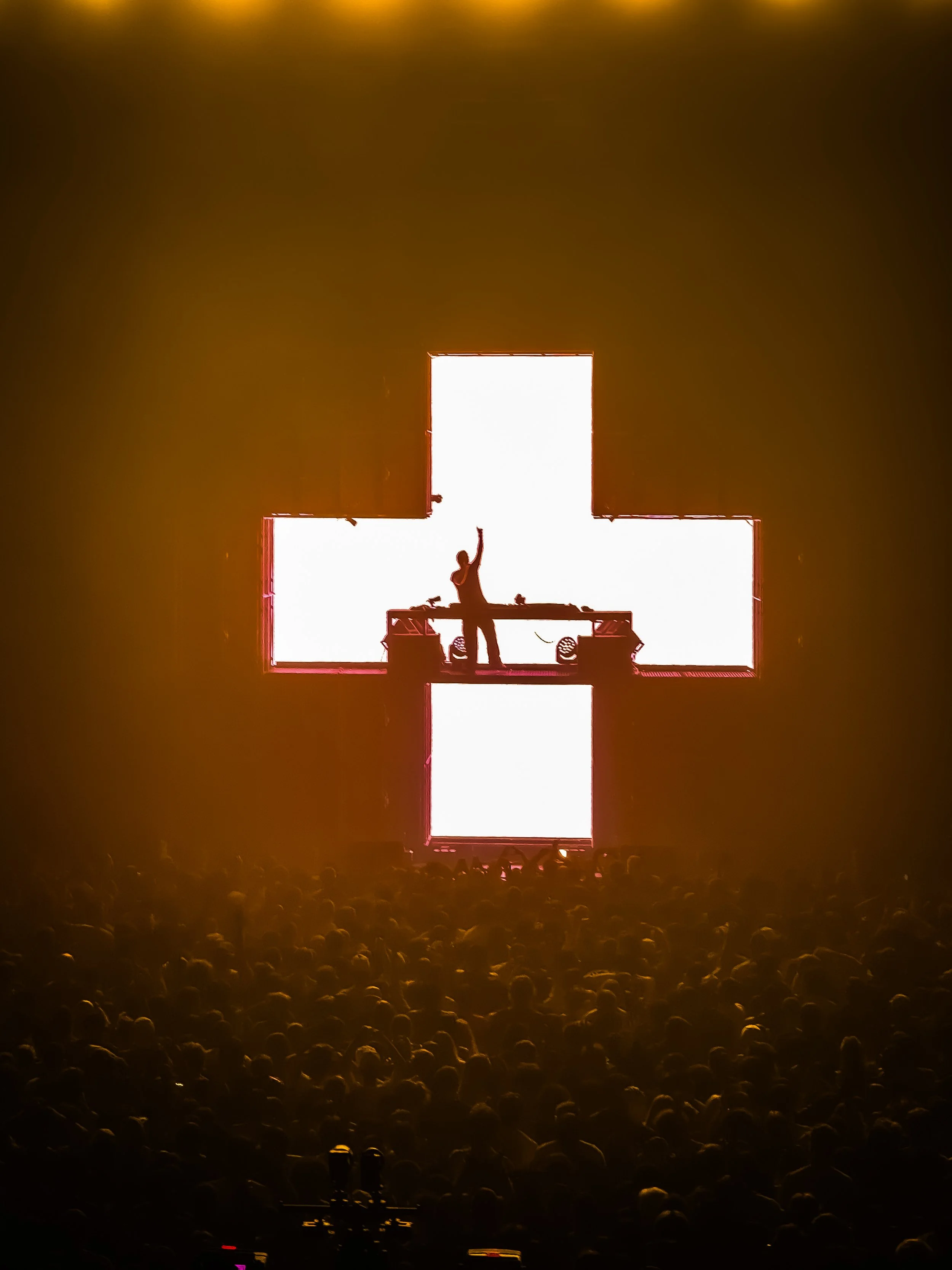 A DJ performs on a stage with a large cross-shaped light display behind him, silhouetted against bright white lights, with a large audience in front.
