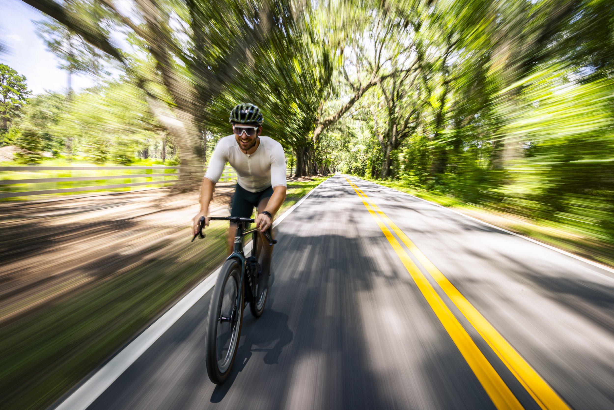 A man wearing sunglasses, a bike helmet, and a white shirt is riding a bicycle on a tree-lined road with yellow dividing lines. The background shows a motion blur of green trees and the road, indicating high speed.