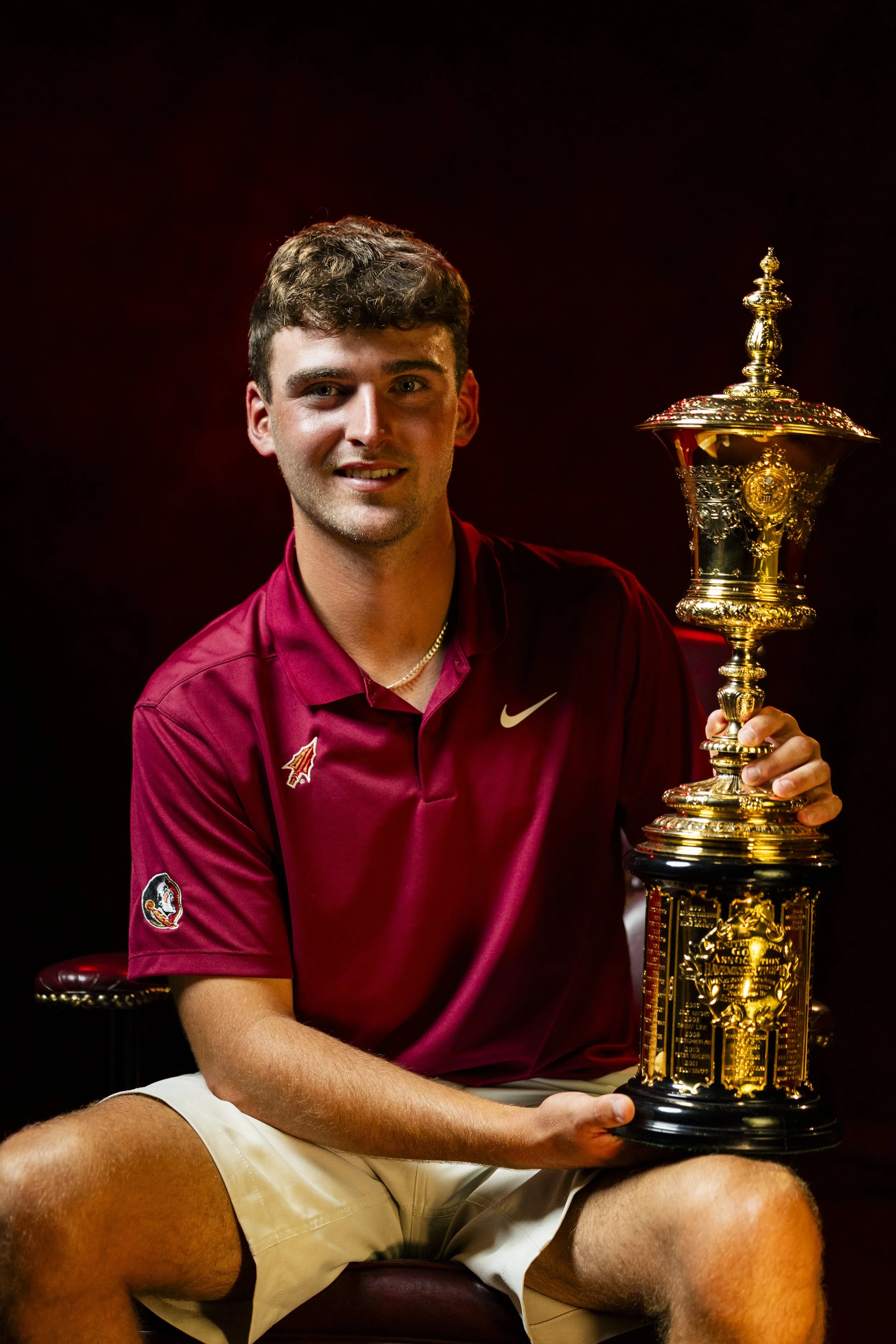 A young man sitting on a bench holding a large, ornate gold trophy. He is wearing a maroon shirt with a logo and beige shorts, and smiling at the camera against a dark background.