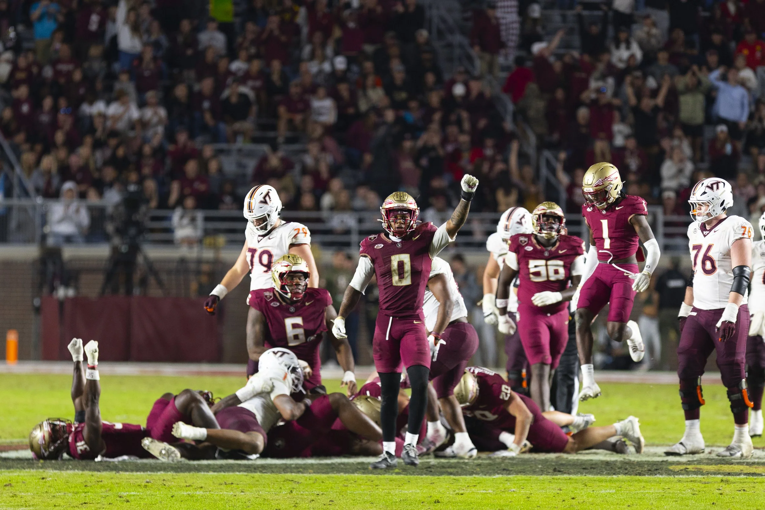 Football players in maroon and white uniforms on the field during a game, with one player raising his fist and others standing or lying on the ground, amid a crowd of spectators.