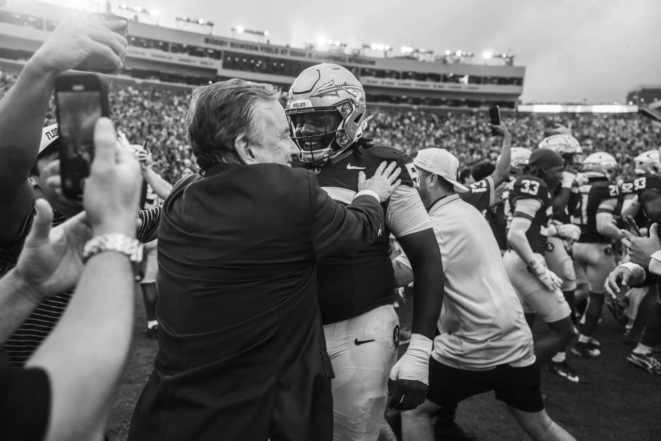 A black and white photo at a football stadium shows a man in a suit hugging a football player wearing a helmet and uniform. Fans are around them, some taking photos, and the stadium is filled with people in the background.