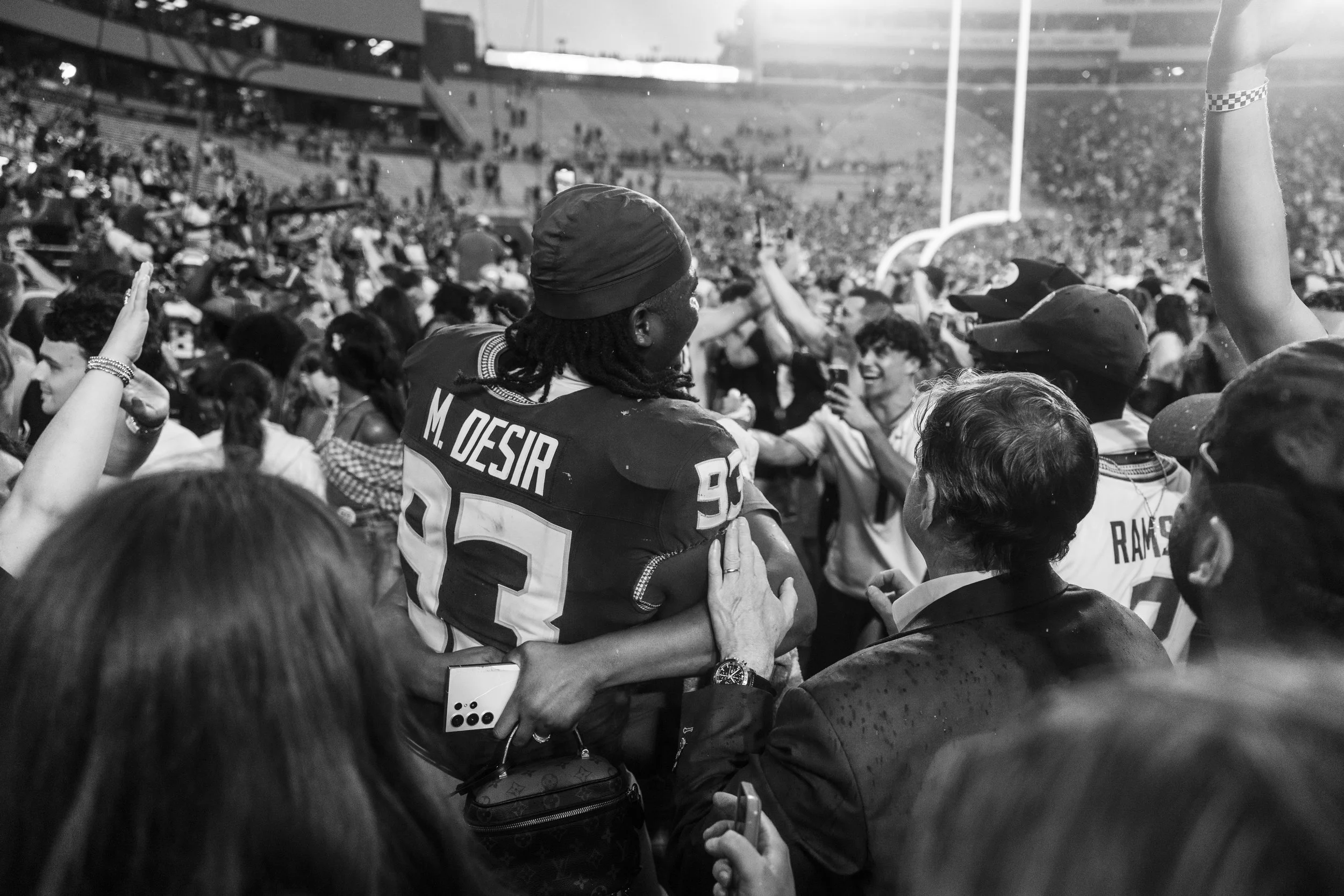 A football player in a jersey with the name 'M. Desir' and the number '25' is being lifted by a woman and is surrounded by a crowd. They are celebrating on a football field with goalposts visible.