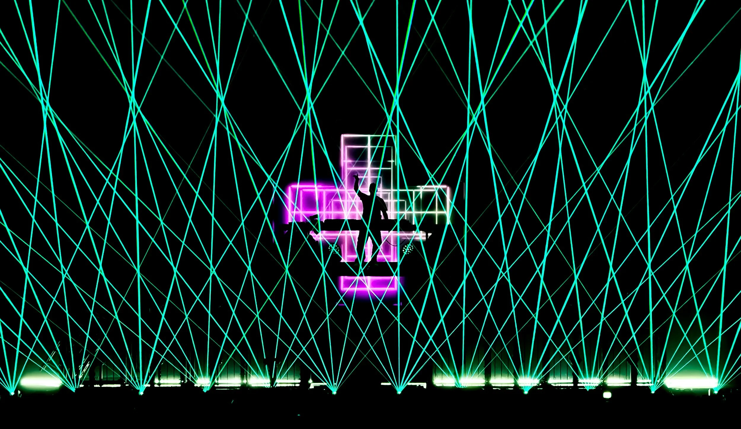 A silhouette of a person appears to be balancing or dancing on a platform illuminated in neon purple, set against a backdrop of neon blue and green laser light beams crisscrossing a black background.