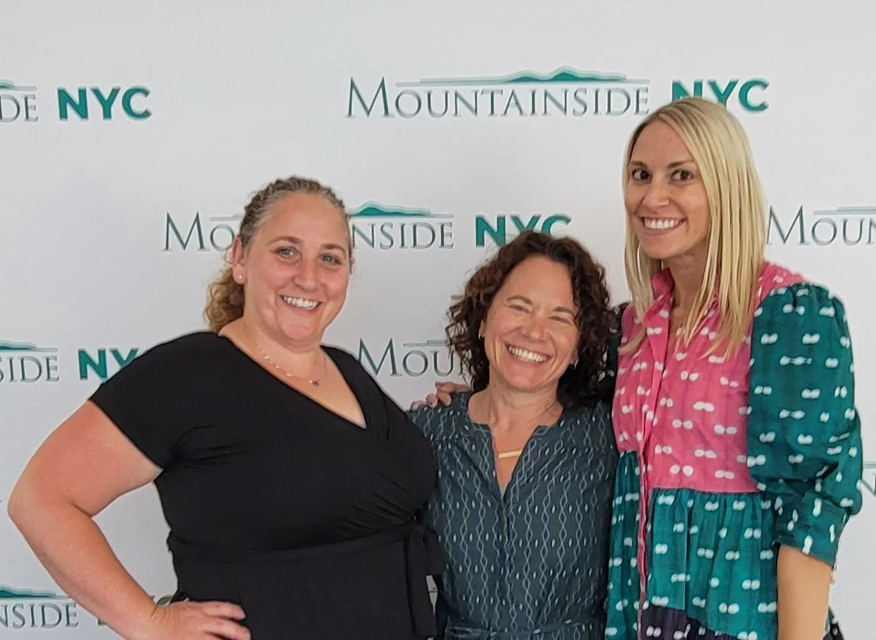 Three women smiling in front of a Mountsideside NYC backdrop, standing close together.