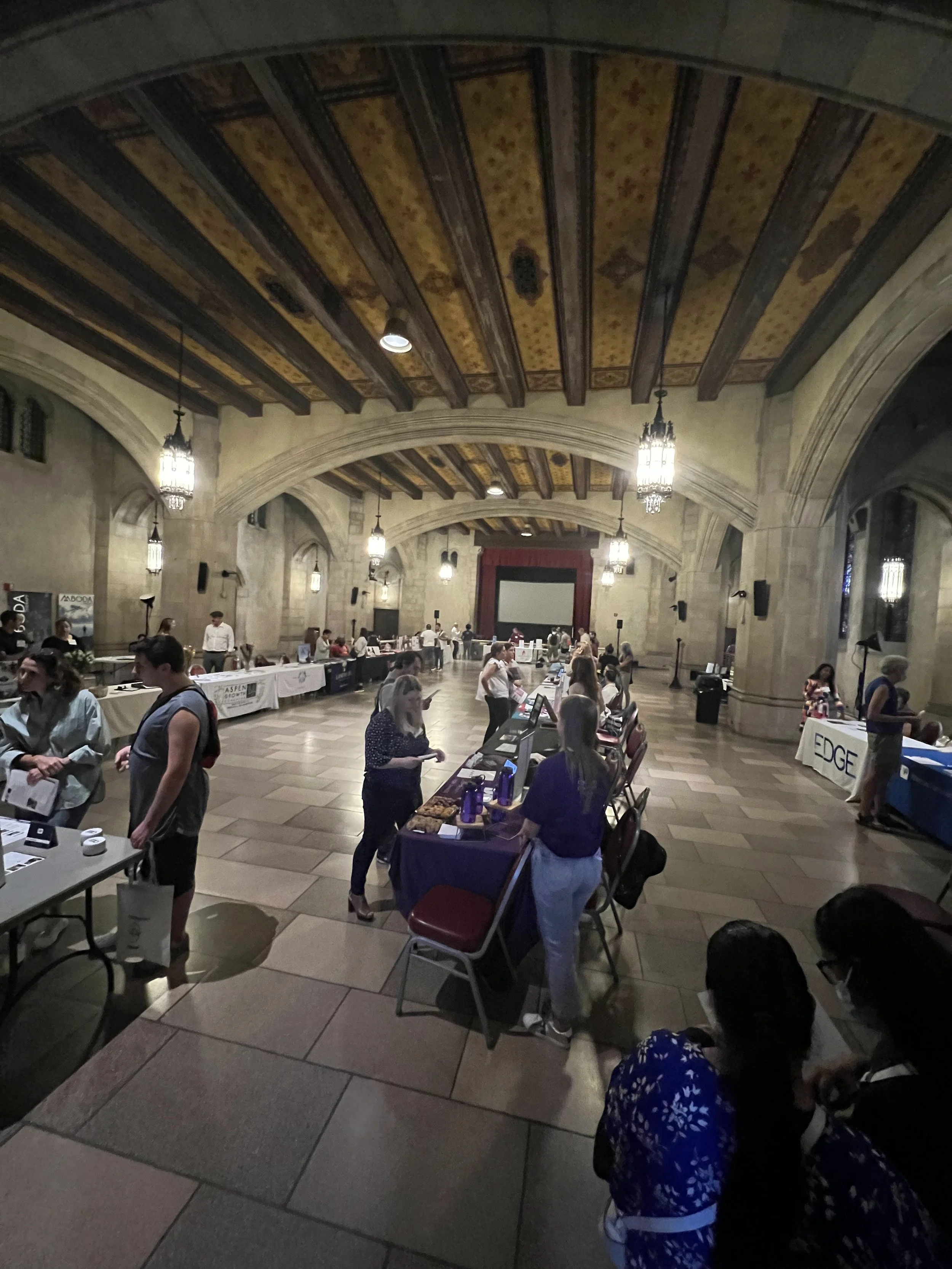 A large, historic hall with a high, ornate wooden ceiling and stone walls, hosting an event with tables and people browsing exhibits.