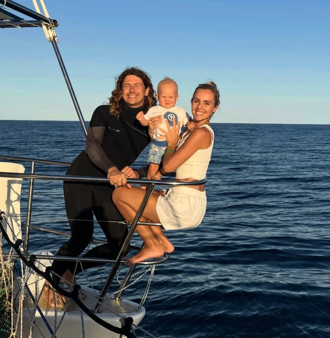 A family of three enjoying time on a sailboat, with the ocean in the background during sunset, smiling and happy.