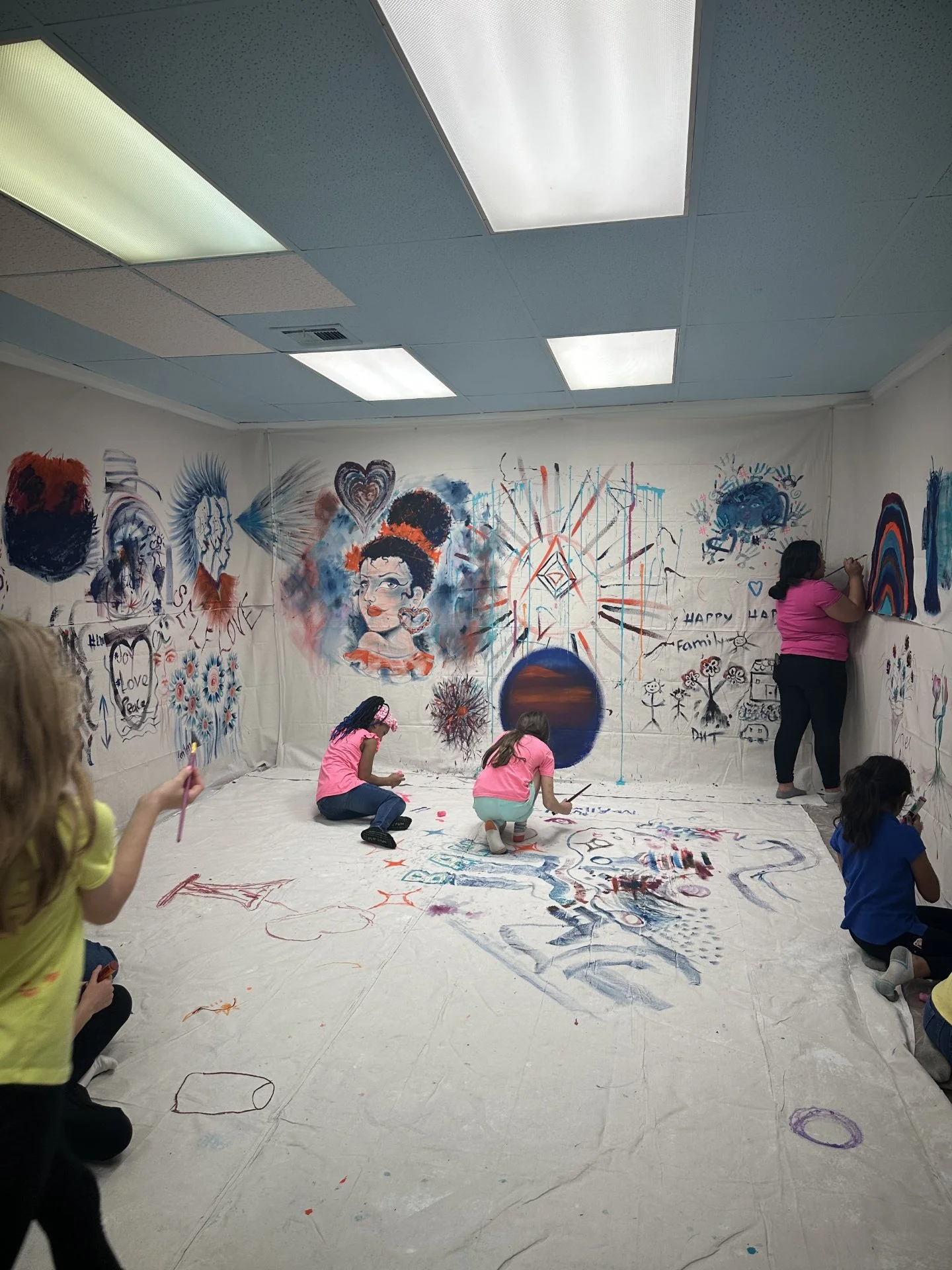 Children painting and drawing on canvas-covered walls in an art room with vibrant, abstract artwork, including portraits, hearts, and various colorful designs.
