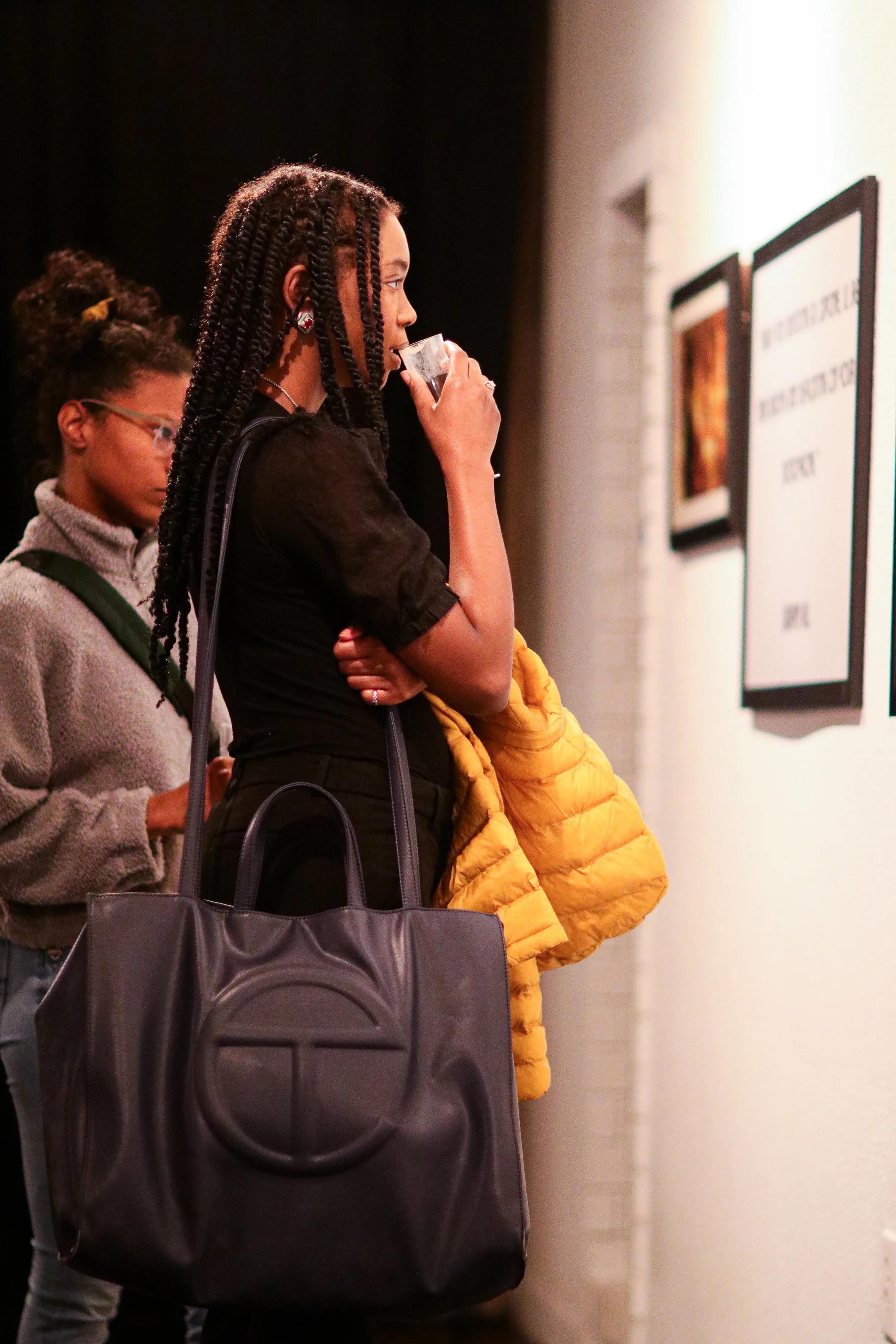 Two people looking at art on a wall in a gallery, with one person holding a drink and wearing a large black bag.