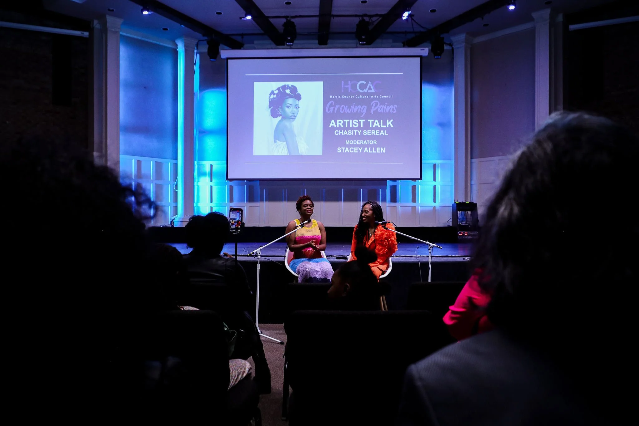 Indoor artist talk event with two speakers seated on stage, audience in the foreground, and presentation screen displaying 'Growing Pains Artist Talk with Chasity Sereal, Moderator Stacey Allen' at the Harris County Cultural Arts Council venue.