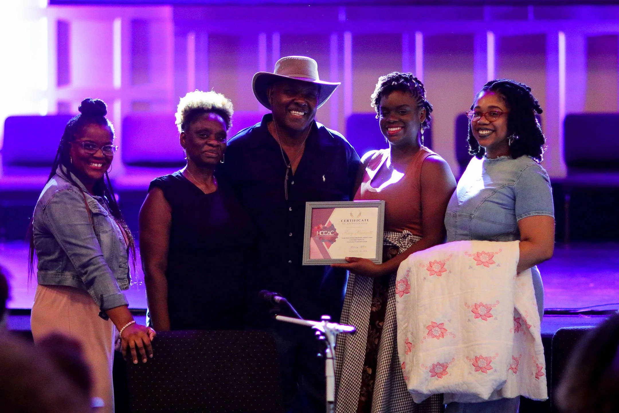 Group of five people posing together; one holds a certificate while another holds a quilt, under stage lighting.