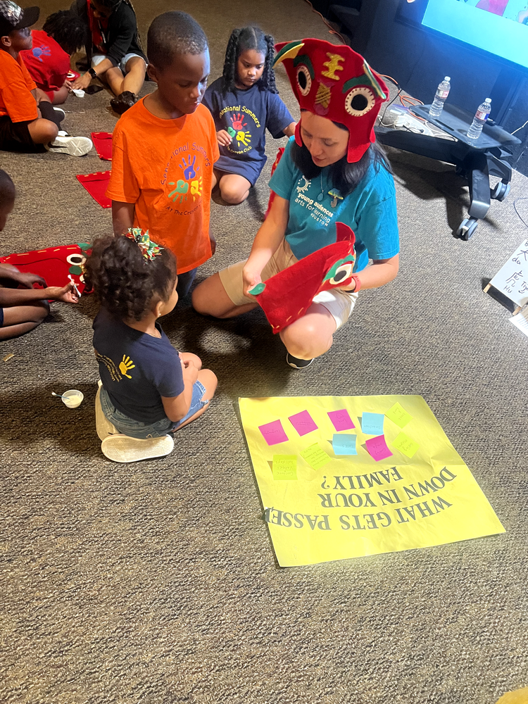 A group of children and an adult sitting on the floor engaged in a craft activity. The adult is holding a red felt piece resembling an animal. A poster on the floor has colorful sticky notes on it, and the text "What gets passed down in your family?" is visible. Several children are wearing colorful t-shirts with artistic designs.
