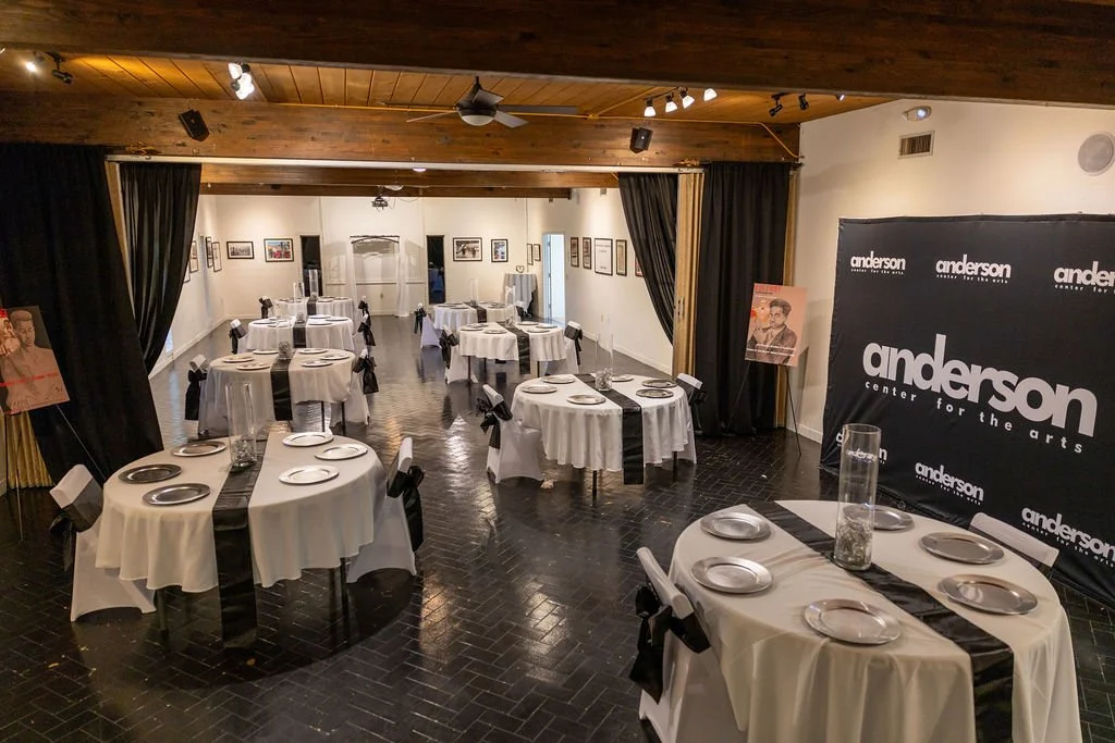 A banquet hall set up with round tables covered in white tablecloths and black table runners, each with silver plates and black bows on chairs. The room features art on the walls and a banner reading "anderson center for the arts."