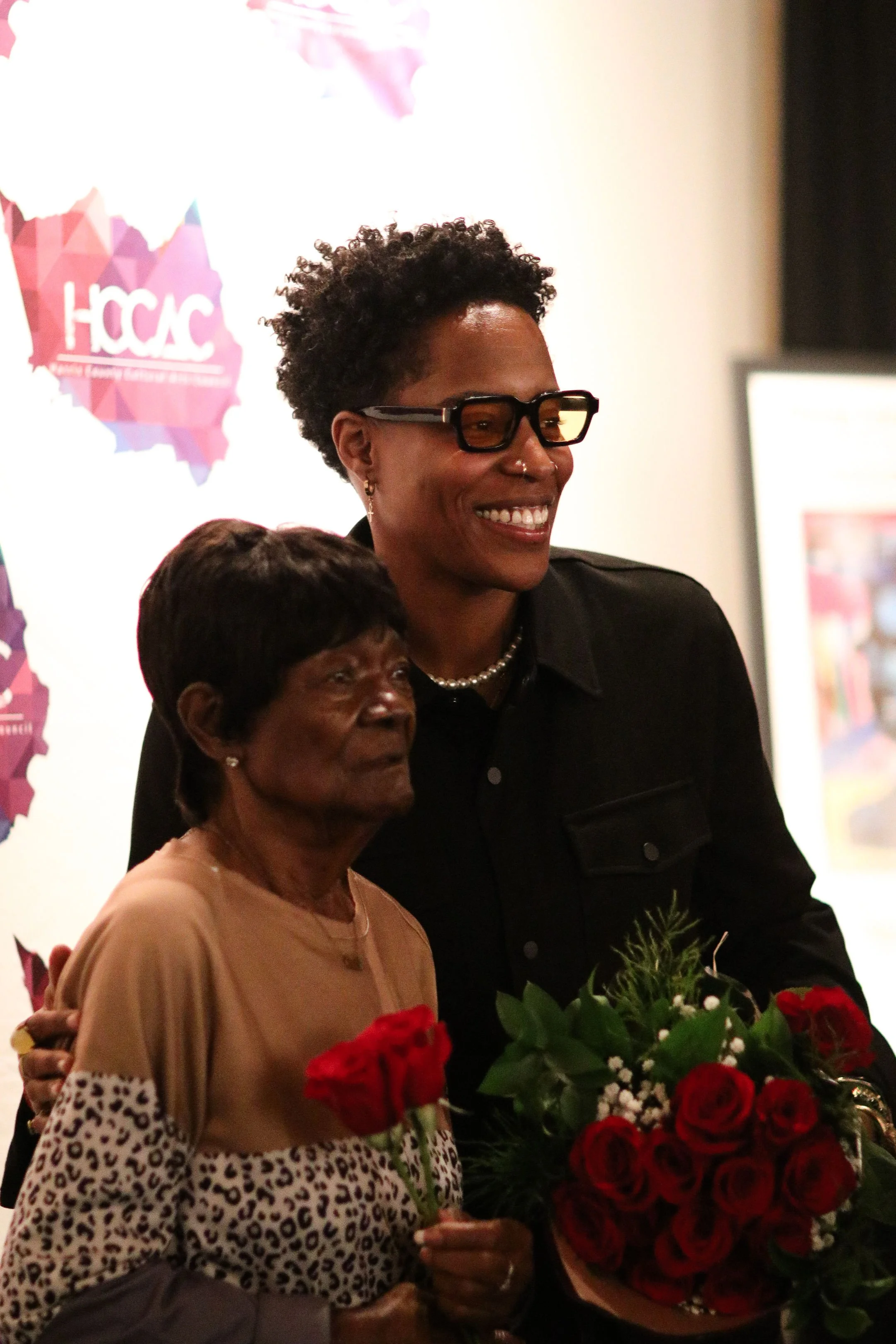 Two smiling people holding red roses at an event; one person wears glasses and a dark jacket, the other wears a leopard-print top. The backdrop displays the logo "HCCAC" in colorful text.