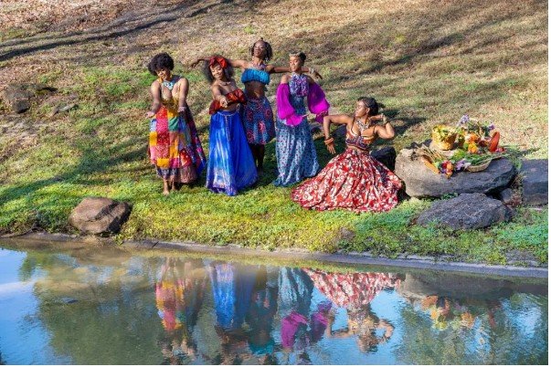 Six women in colorful dresses pose by a pond with reflections in the water.
