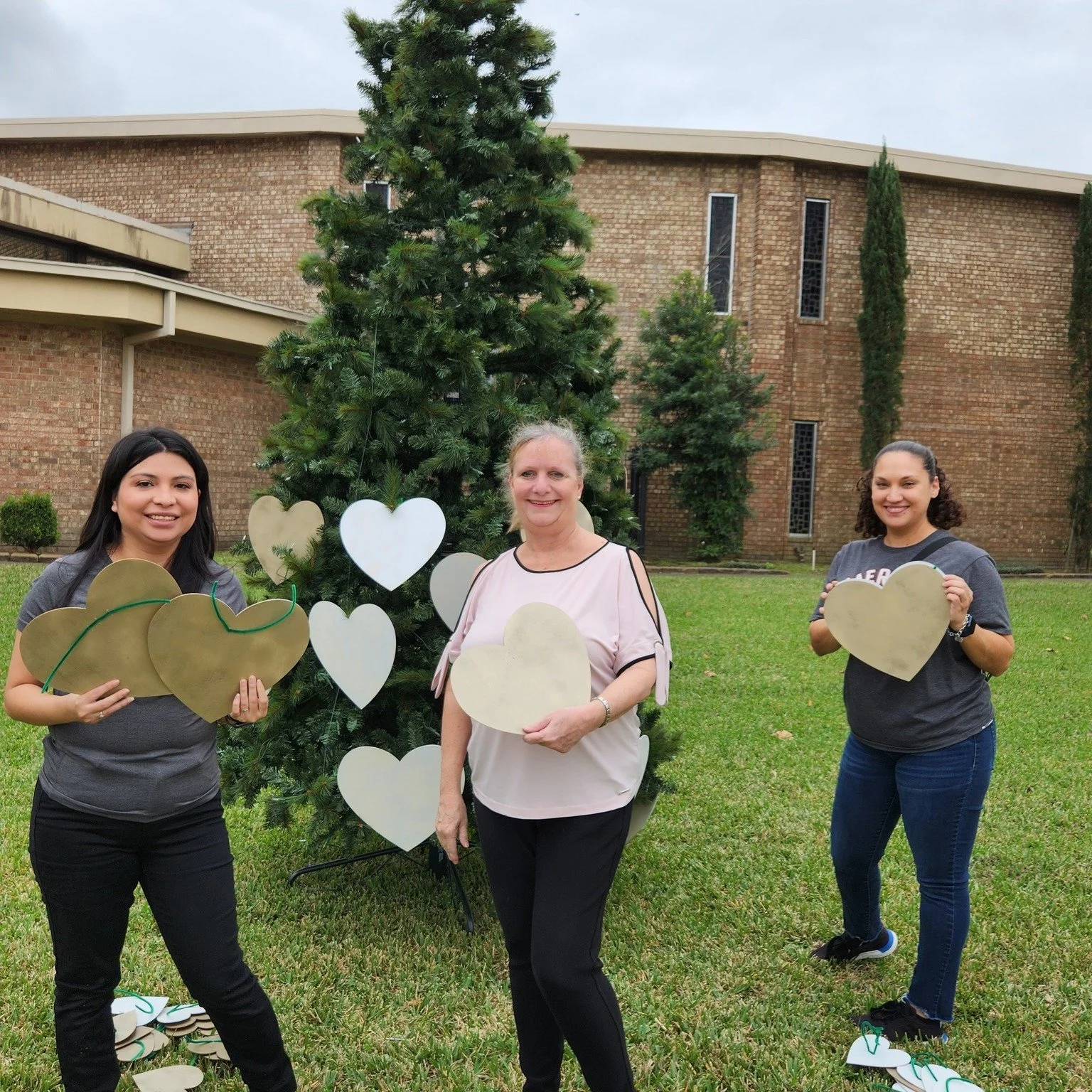 Three women standing on grass holding heart-shaped cutouts in front of a tall decorated tree with more heart-shaped ornaments, outside a brick building.