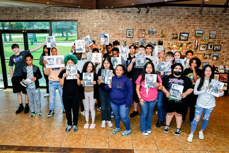 A diverse group of students standing indoors holding black and white photos, with a brick wall and framed pictures in the background.
