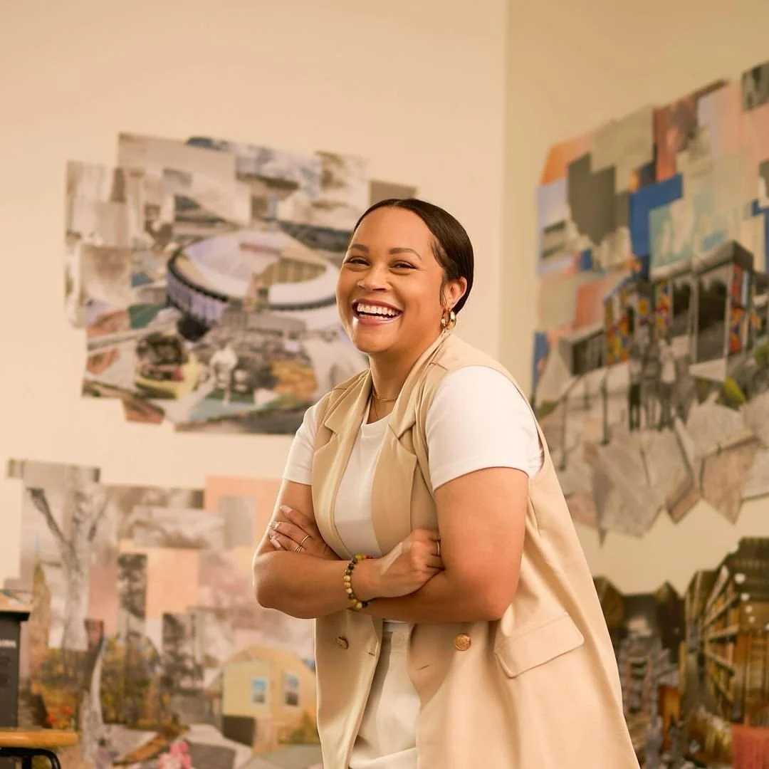 A person smiling and standing with arms crossed in front of a wall displaying abstract art or architectural designs.