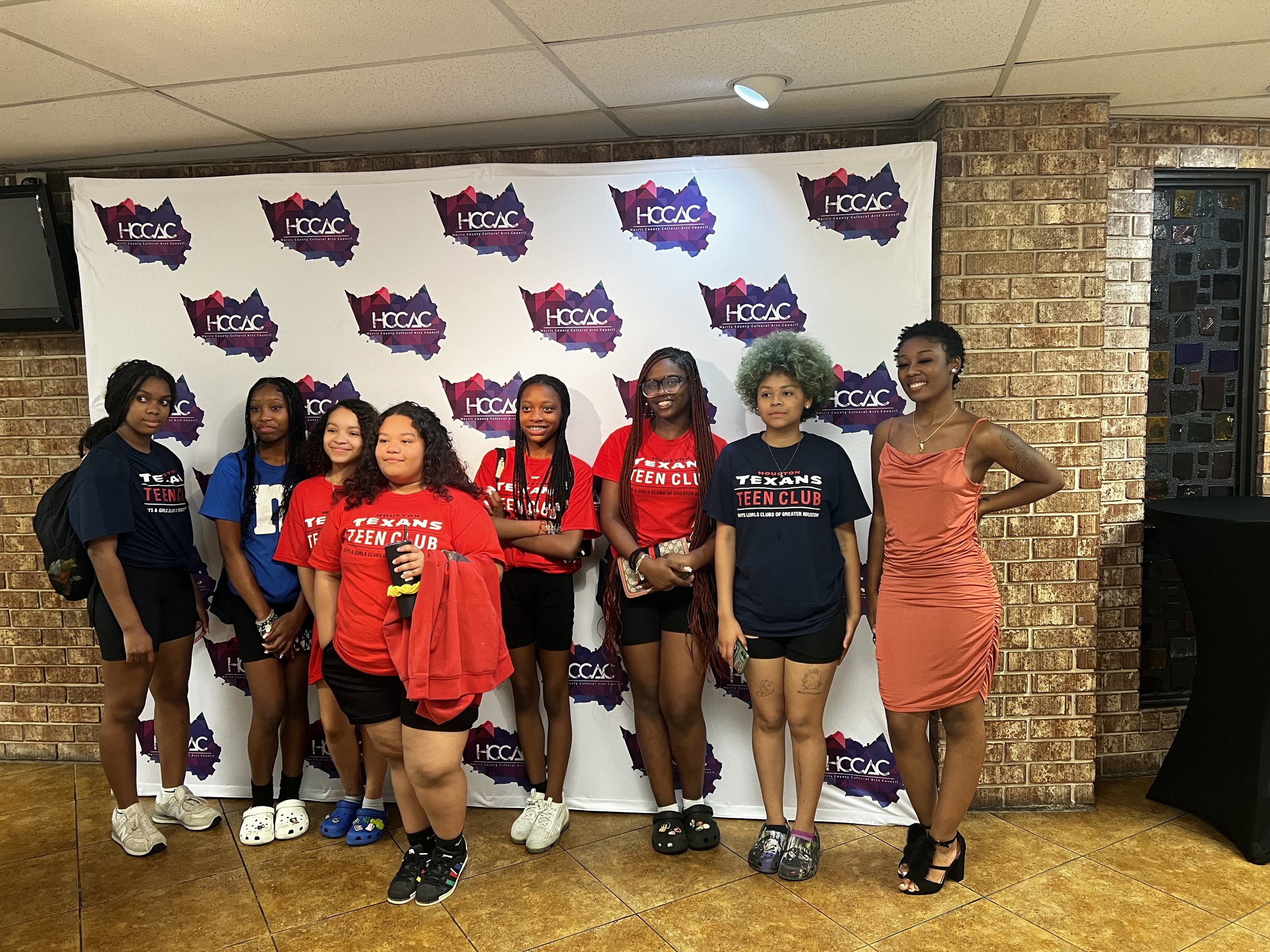 Group of people posing in front of a backdrop with HCCAC logo, wearing casual clothes and Texans Teen Club shirts.