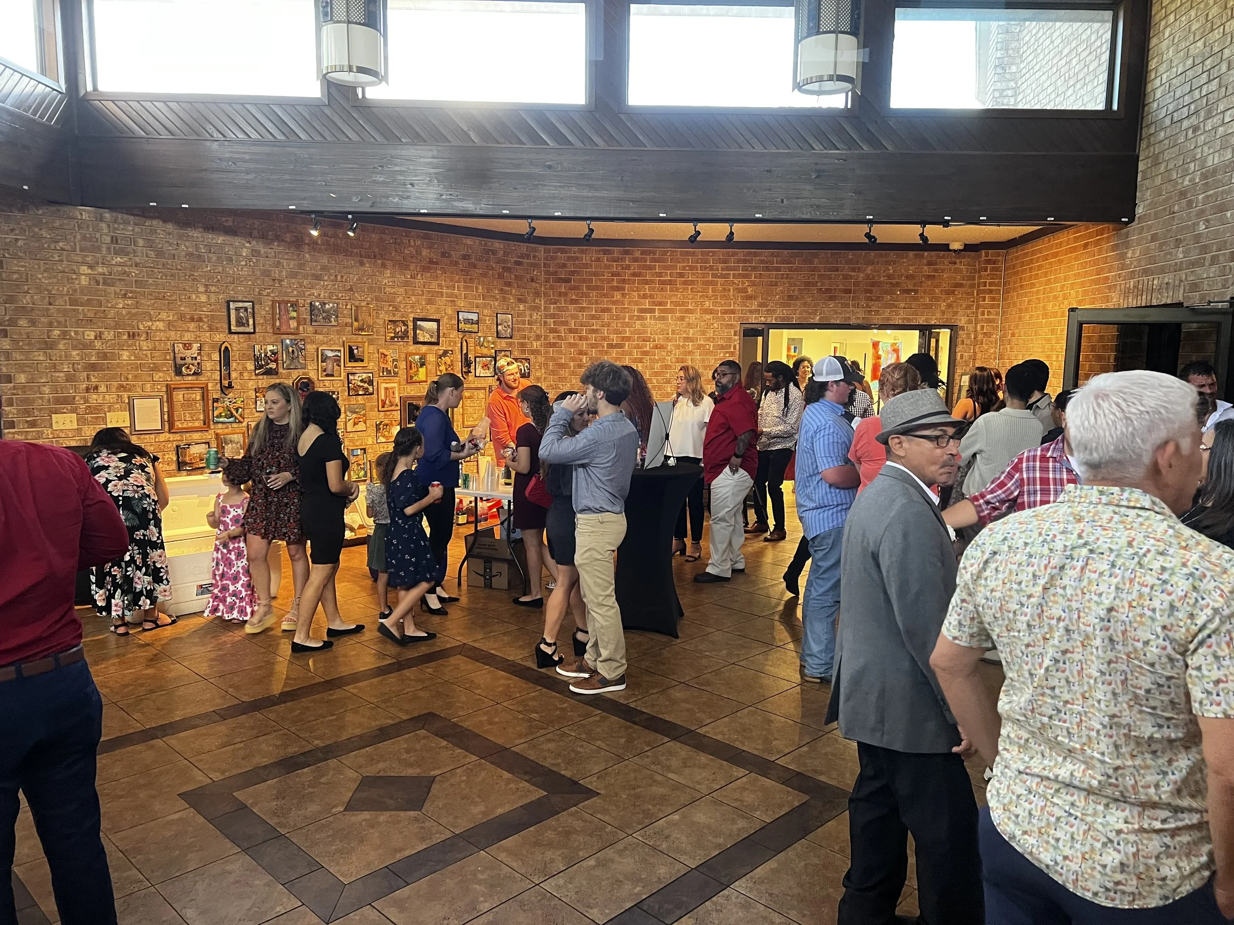A crowded indoor gathering with people socializing, featuring a brick wall decorated with pictures and artwork. Guests are wearing casual and semi-formal attire, mingling and engaging in conversation. The venue has large windows and warm lighting.