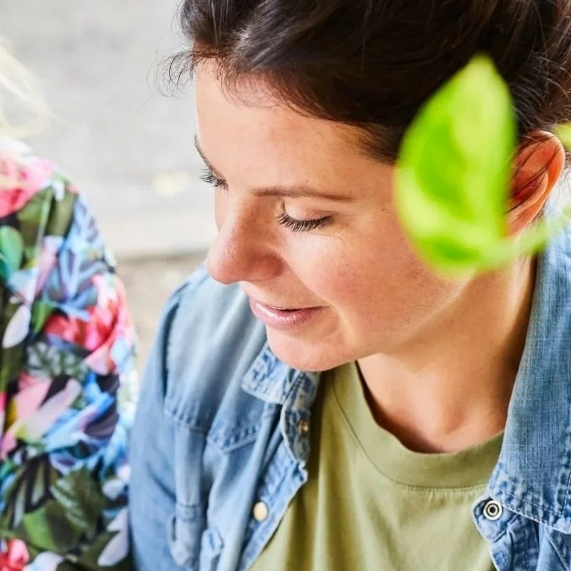 A woman with closed eyes smiling, wearing a denim jacket and a green shirt, next to a person in a colorful floral top, outdoors with a blurred background.