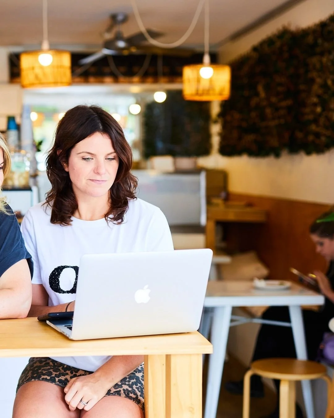 Woman with shoulder-length brown hair sitting at a wooden table working on a silver MacBook in a cozy cafe