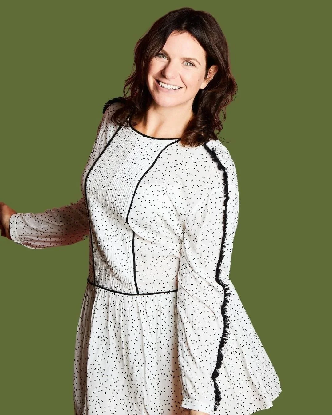 A woman with dark brown hair wearing a long-sleeved white dress with black polka dots and black piping, standing against a solid green background, smiling at the camera.
