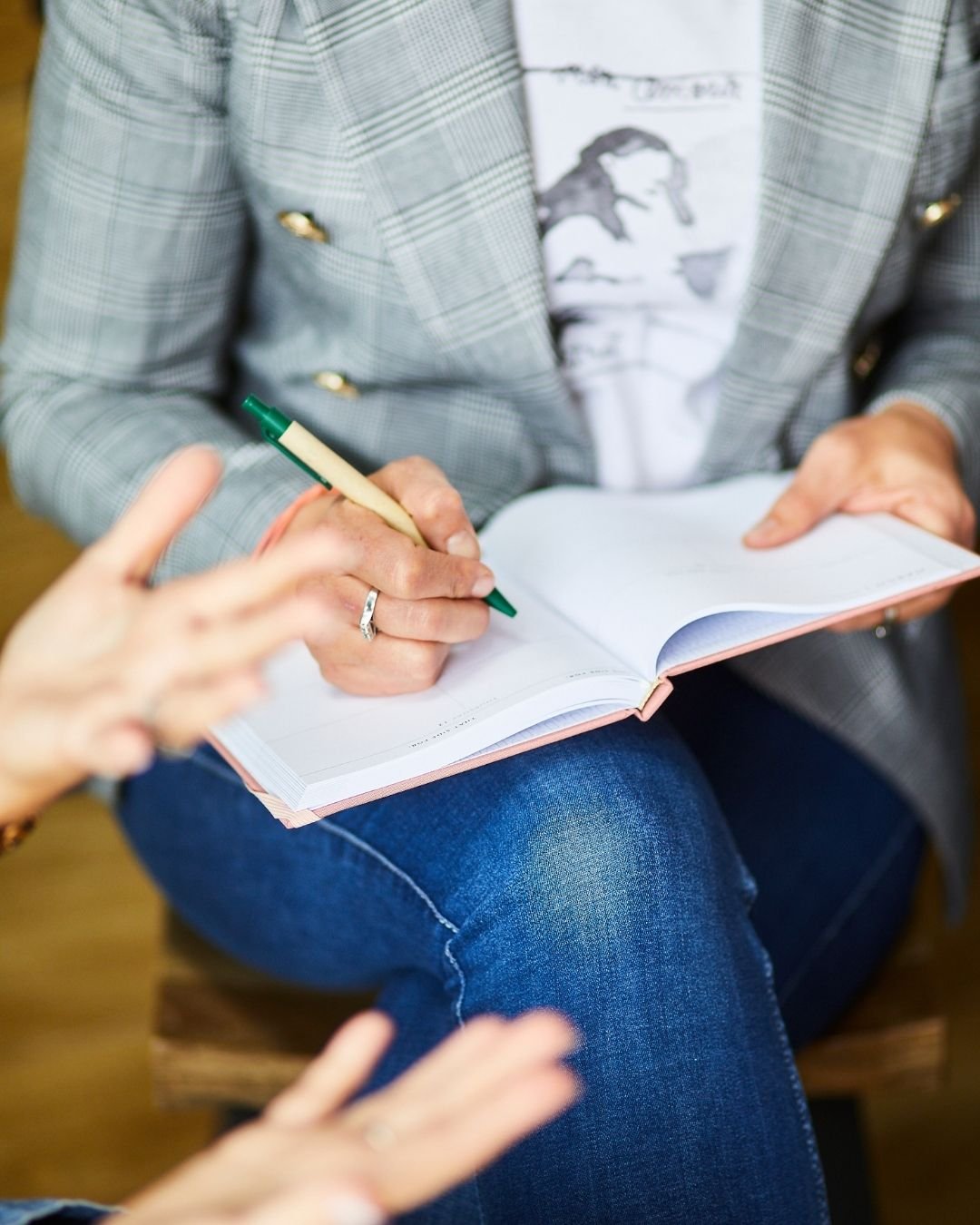 Person wearing a plaid blazer and graphic t-shirt writing notes in a notebook while seated.