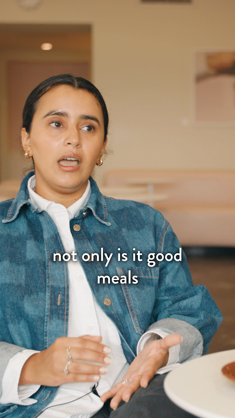A woman wearing a denim jacket and white shirt speaking with subtitle "not only is it good meals". She is seated at a table with a plate of food visible in the foreground.