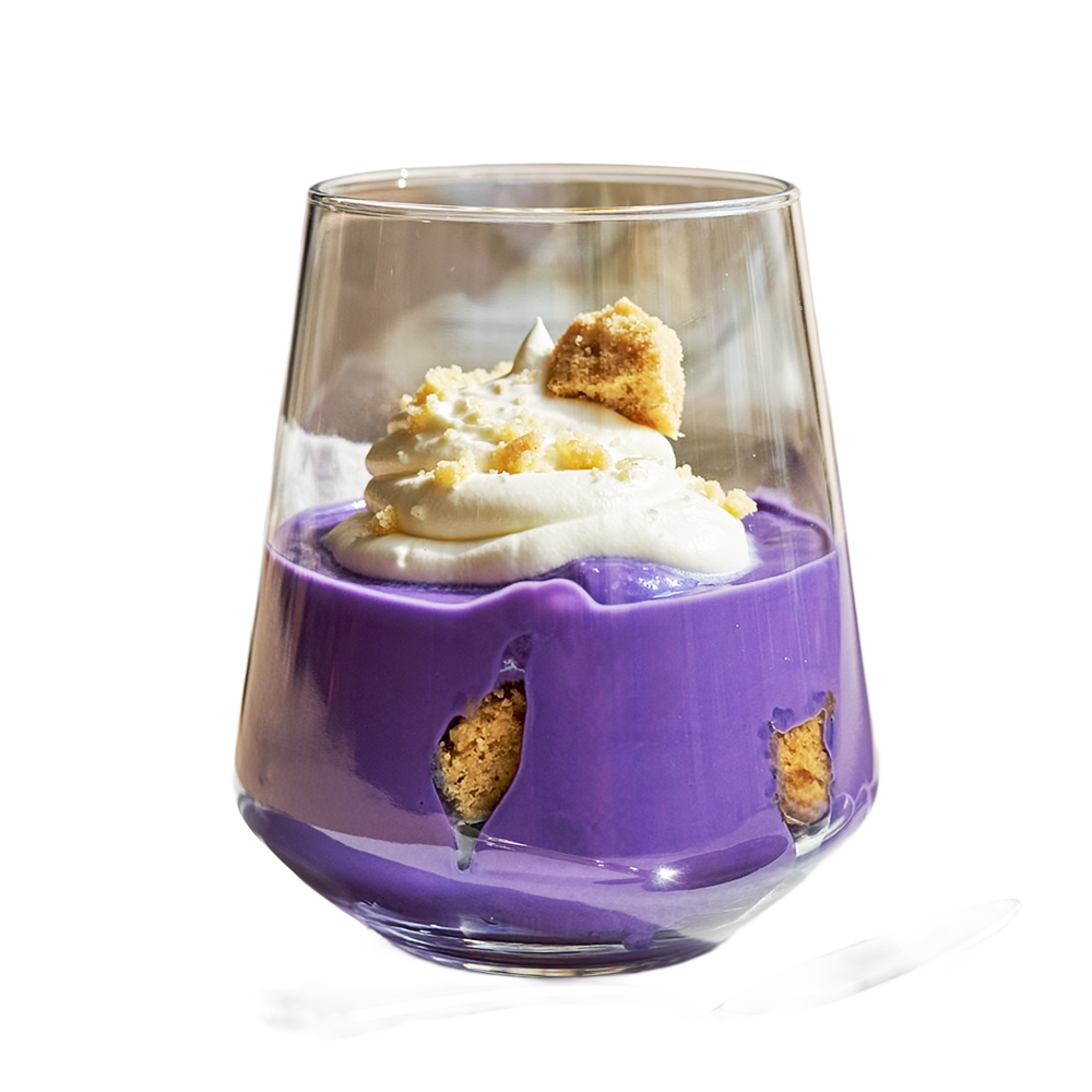 A glass dessert cup filled with purple custard or pudding, topped with whipped cream and cookie crumbles, with cookie pieces visible inside the pudding. A spoon is placed beside the cup.