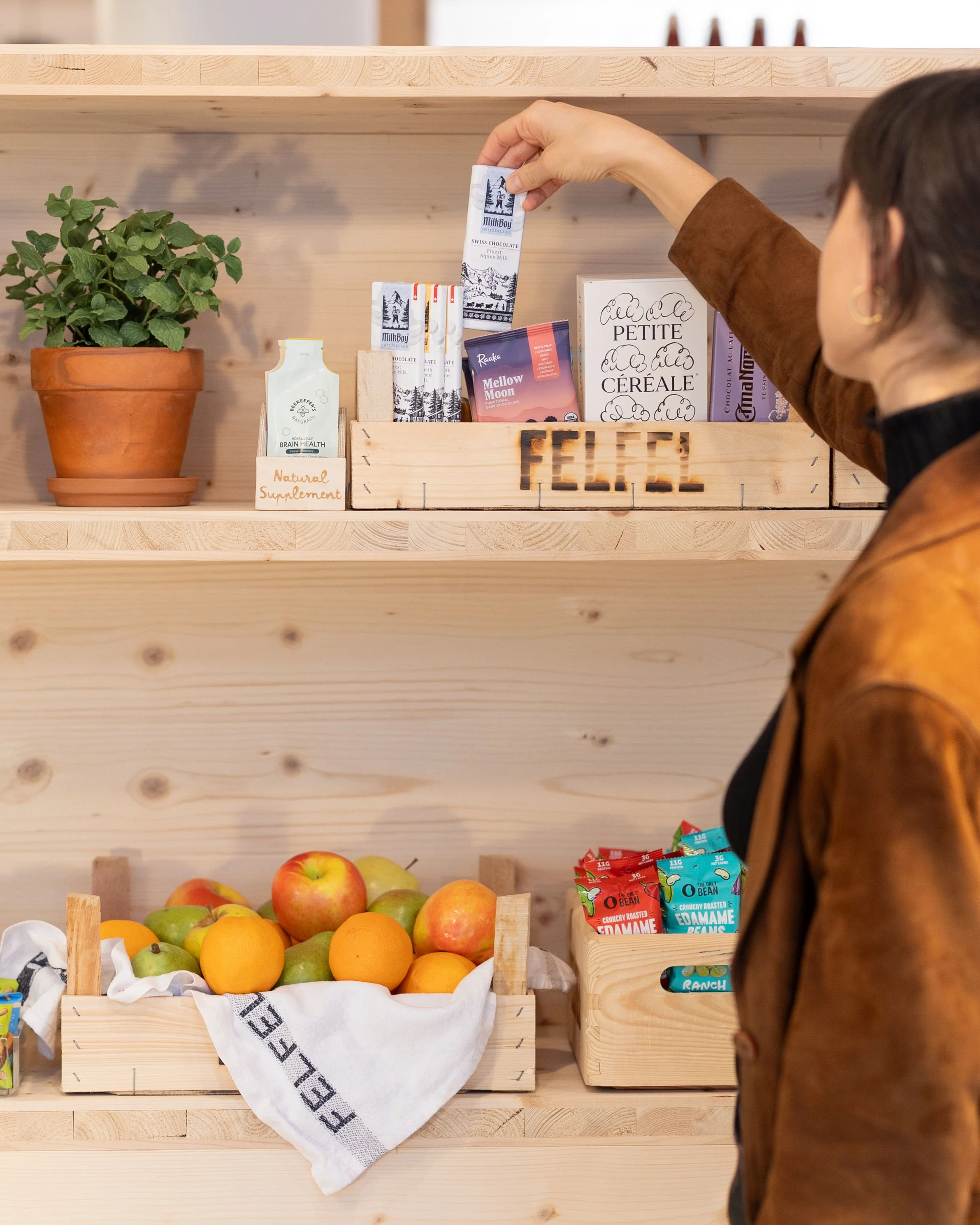 Healthy Office Snacks: A woman looking at a wooden shelf with various products, including a potted plant, boxes of chocolates, a sign, and a crate of apples and oranges.