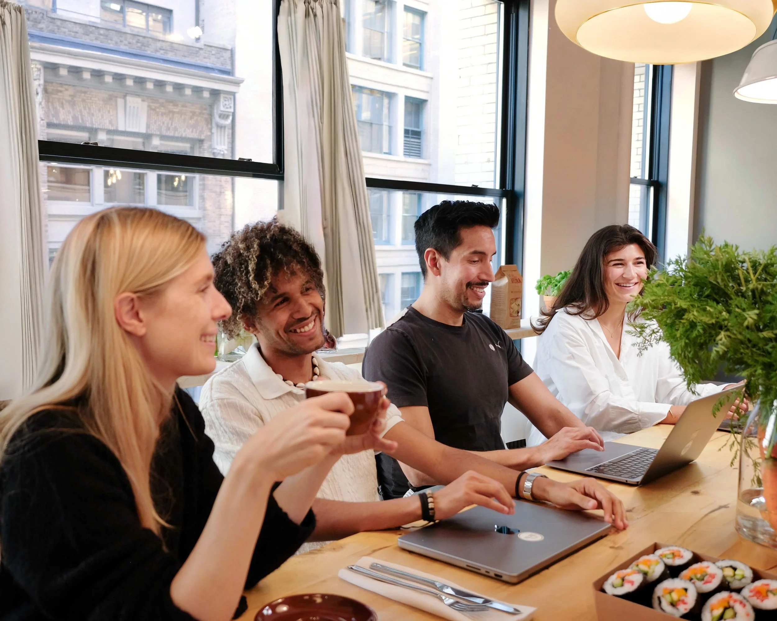 Four people sitting at a dining table with sushi, laptops, and a coffee mug, smiling and talking in a bright room with large windows and curtains.