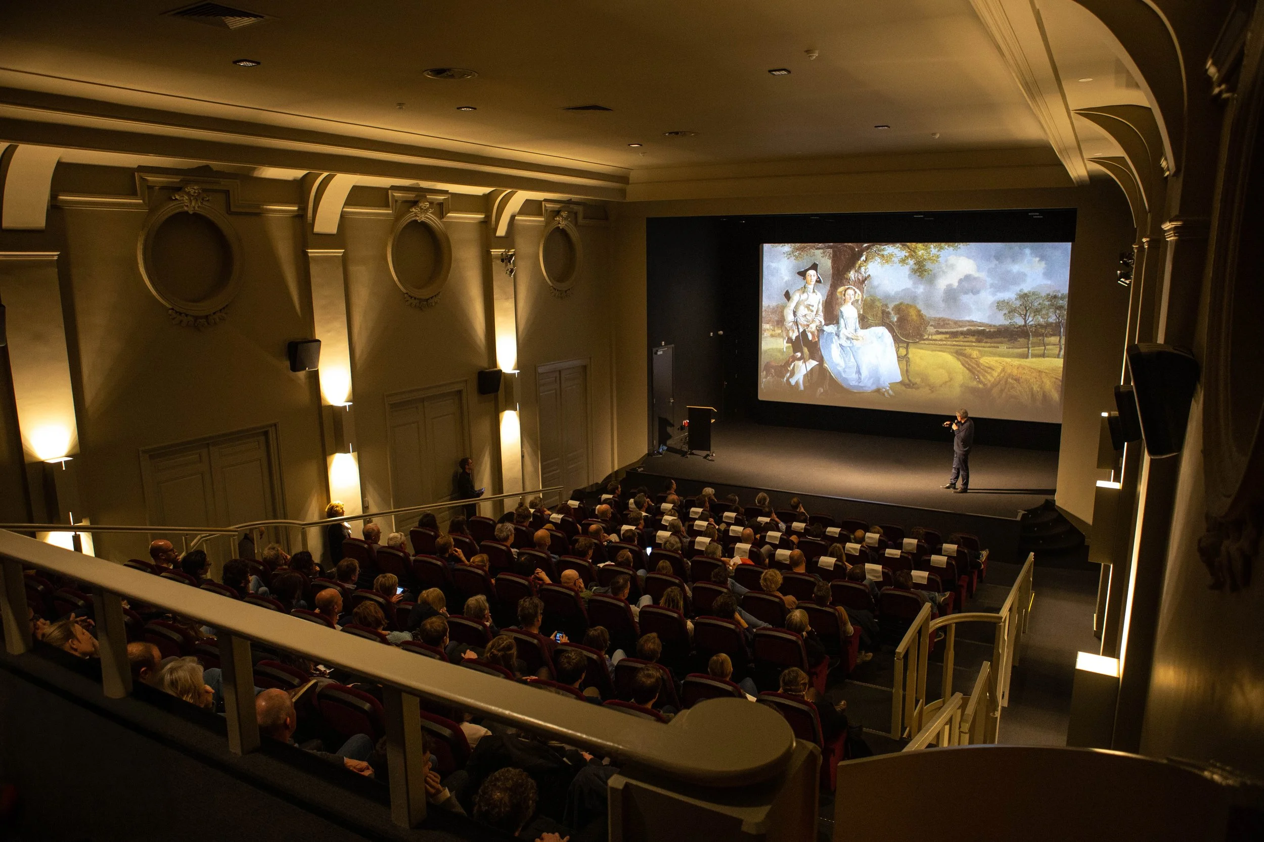 Een groot theater met een publiek dat naar een presentatie kijkt, waarbij een spreker op het podium staat en een groot scherm met een afbeelding van een schilderij toont.