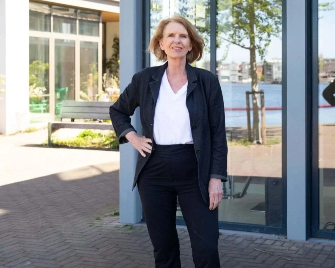 Een vrouw met rood haar, gekleed in een zwarte jas en witte blouse, staat buiten voor een glazen gebouw met uitzicht op een rivier.