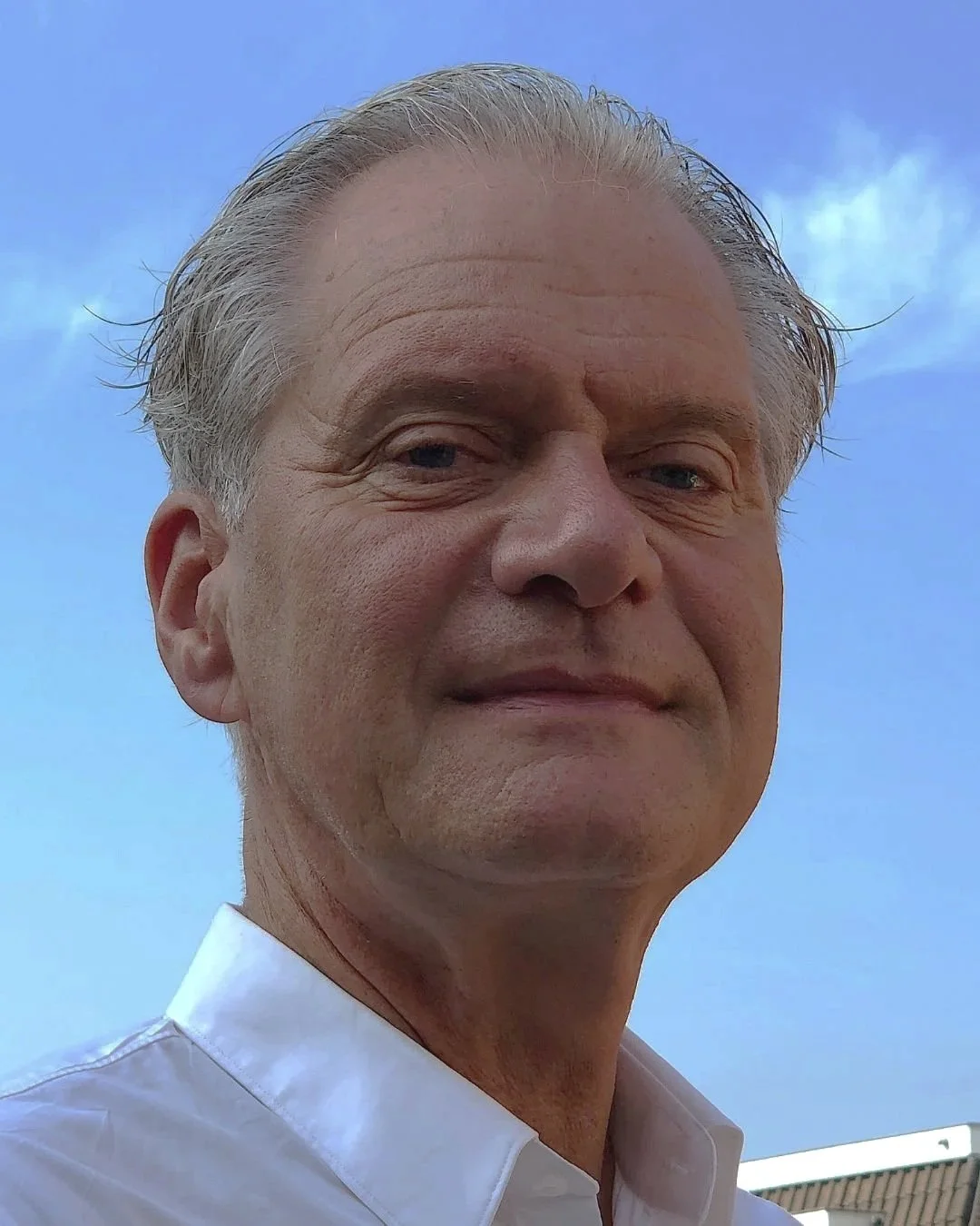 Close-up van een man met grijs haar tegen een blauwe lucht.