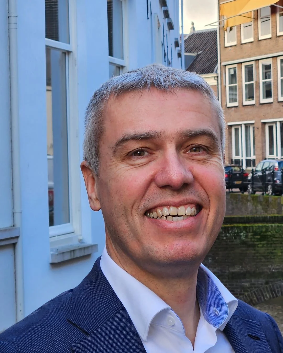 Portret van een lachende man met grijs haar, wit overhemd en donkerblauwe jas, standing buiten in een stad met historische gebouwen.