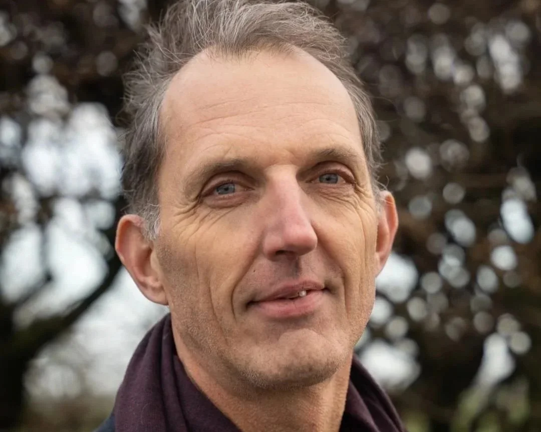 Portret van een man met grijs haar, blauwe ogen en een lichte glimlach, buiten met een achtergrond van bomen en een bewolkte lucht.