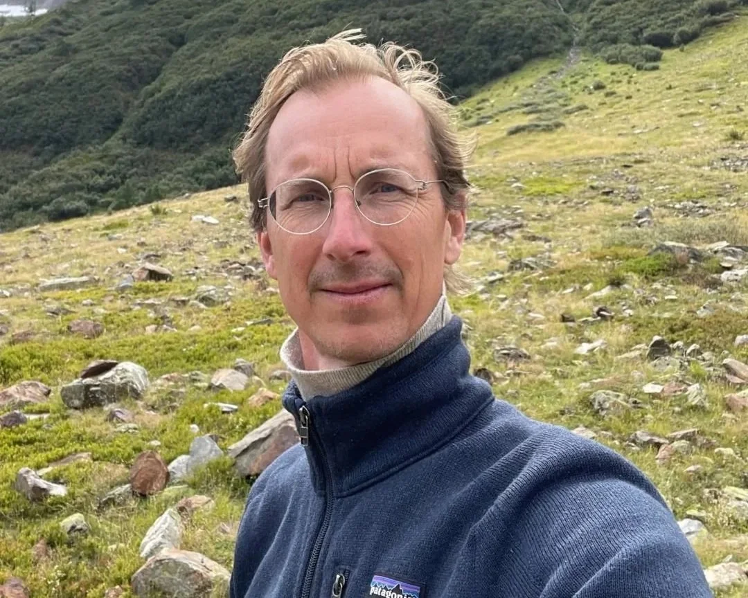 Man met bril en jasje in een groene, bergachtige omgeving met rotsen.